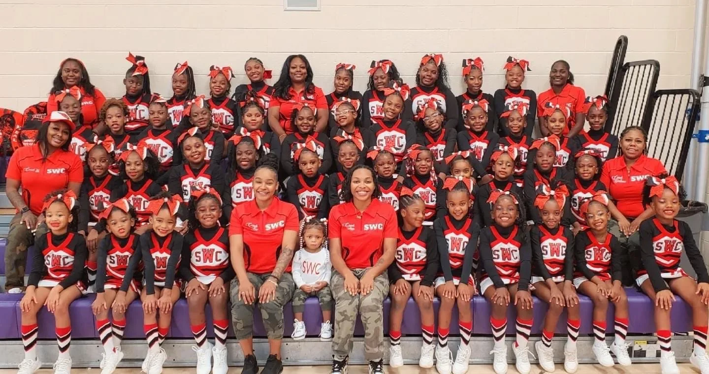 Group of young cheerleaders in matching red, black, and white uniforms posing for a team photo inside gym bleachers, with coaches on either side.