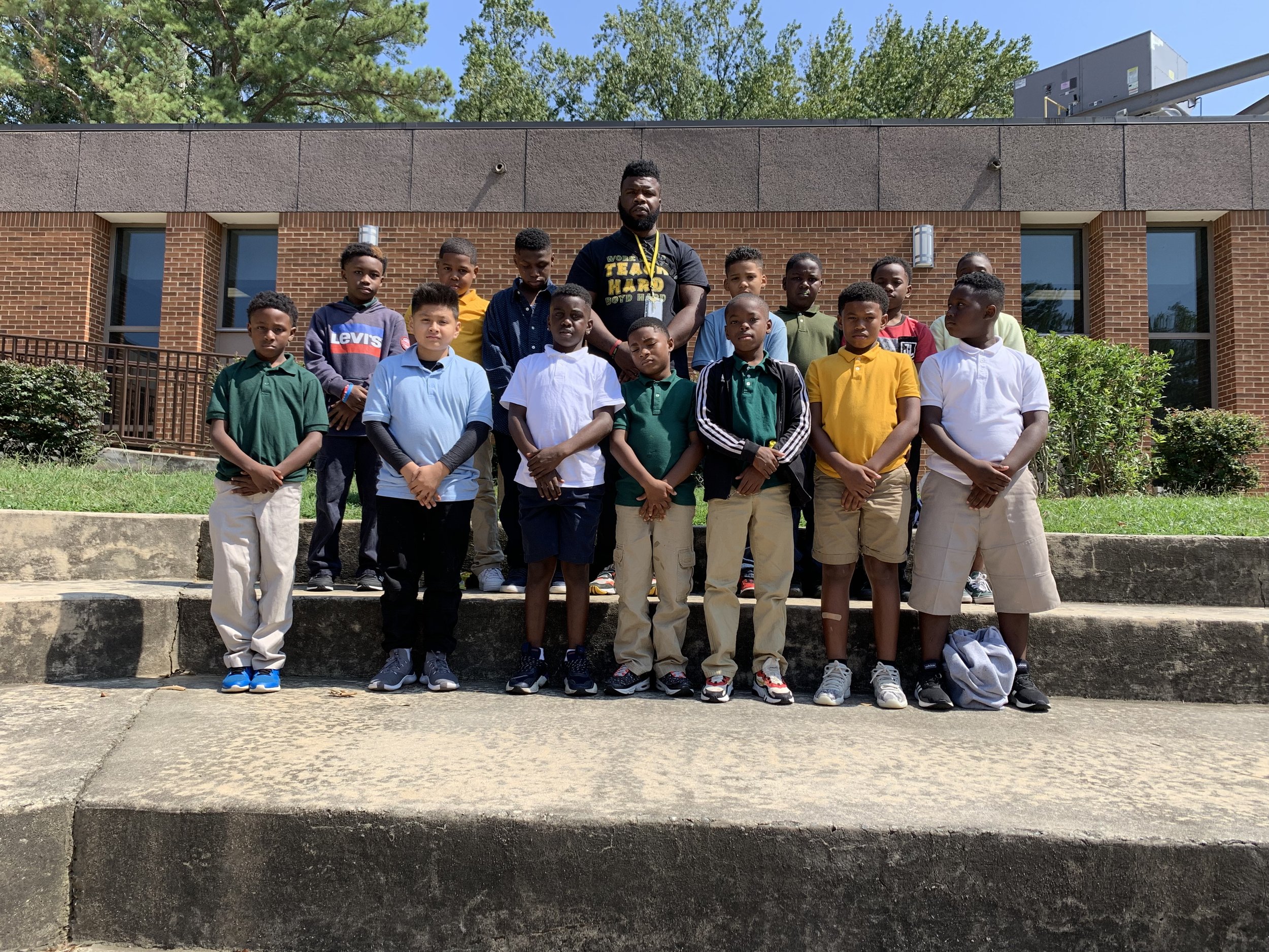 A group of young boys and one adult male standing on outdoor stairs in front of a brick building, posing for a group photo.