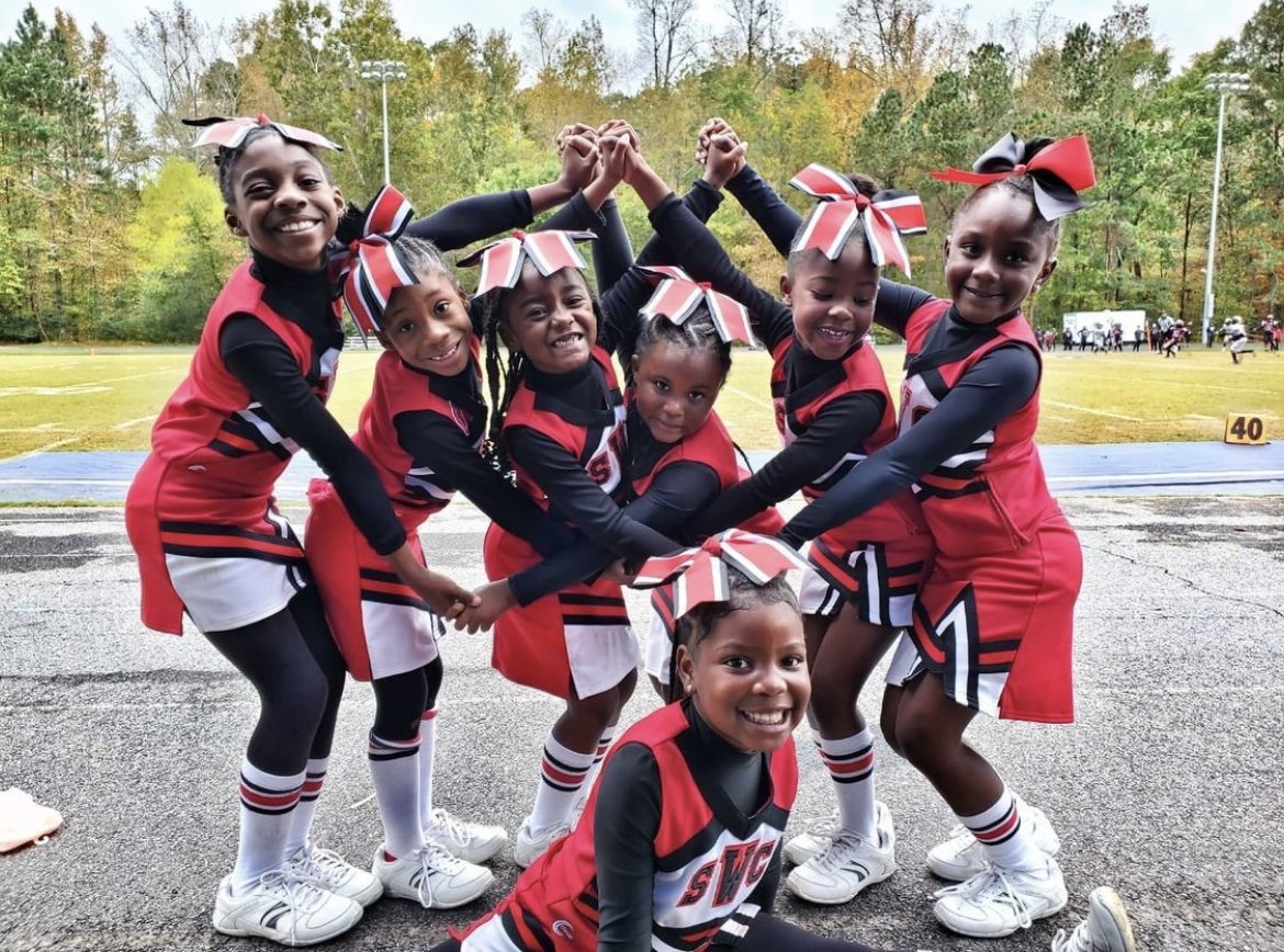 Group of young girls cheerleaders in red and black uniforms with bows in their hair, smiling and posing together outdoors after a game on a track field.