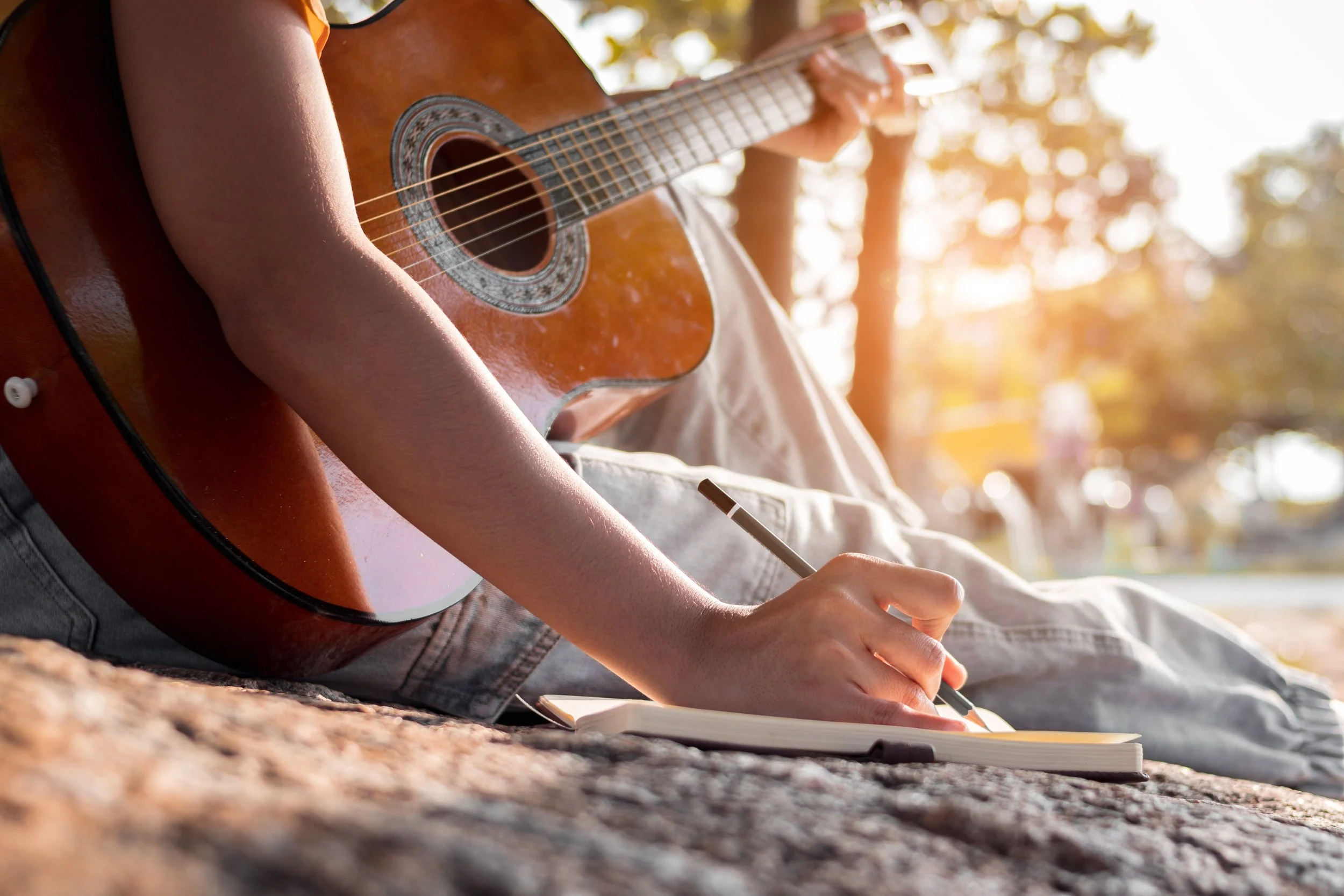 Person sitting outdoors at sunset playing acoustic guitar and writing in a notebook.