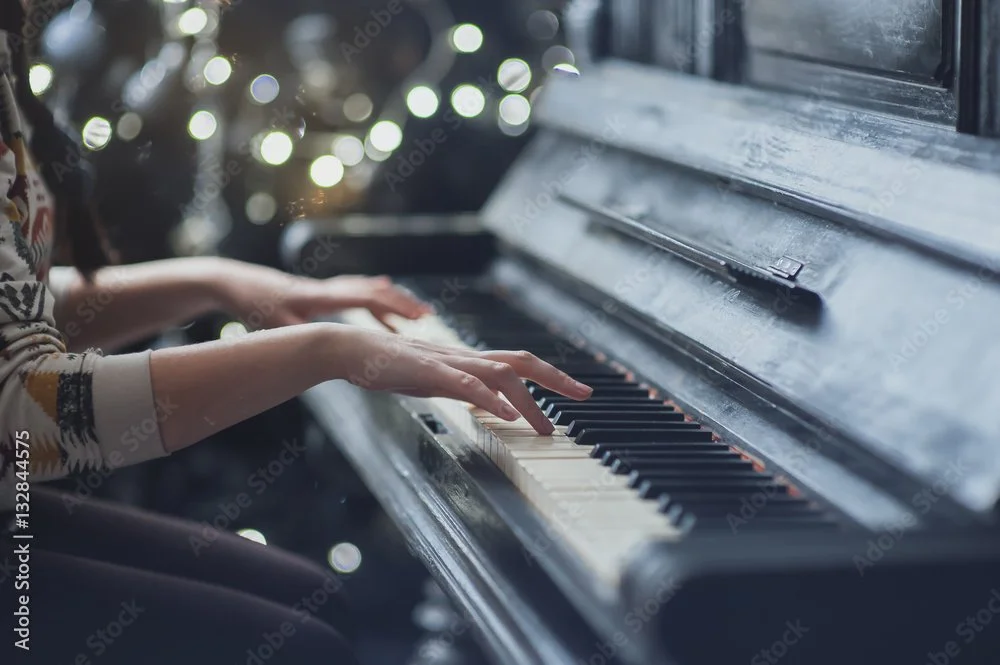 A person playing a black upright piano with a Christmas tree in the background.