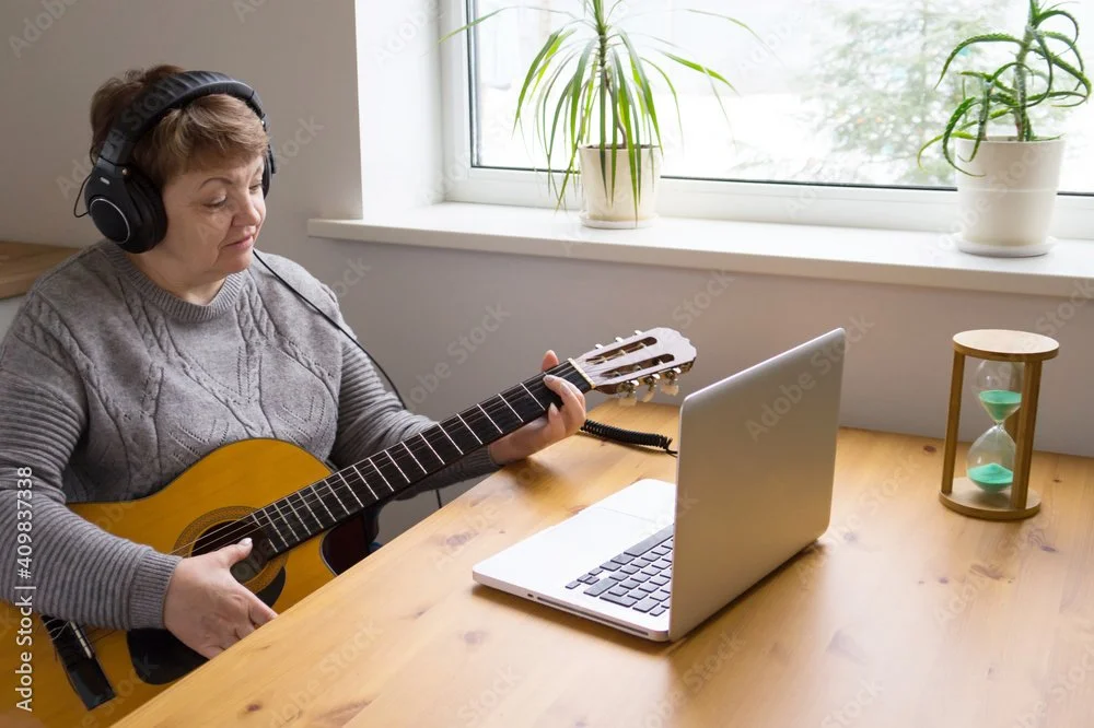Older woman sitting at a wooden table, wearing headphones, playing an acoustic guitar, with a laptop, a potted plant, and an hourglass on the table near a window.