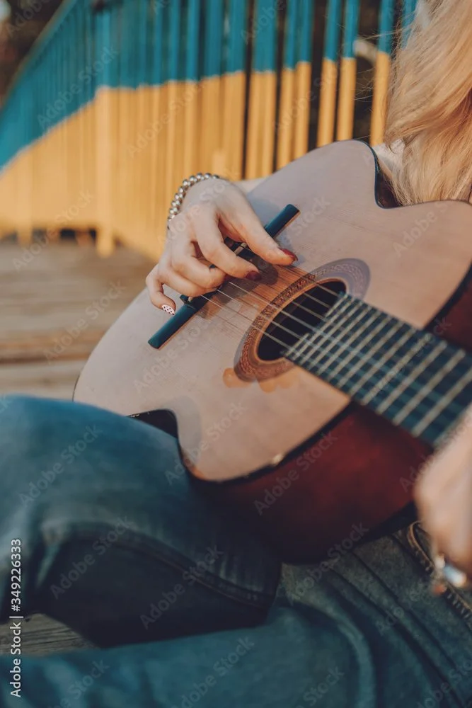 A person playing an acoustic guitar outdoors with a bamboo fence in the background.