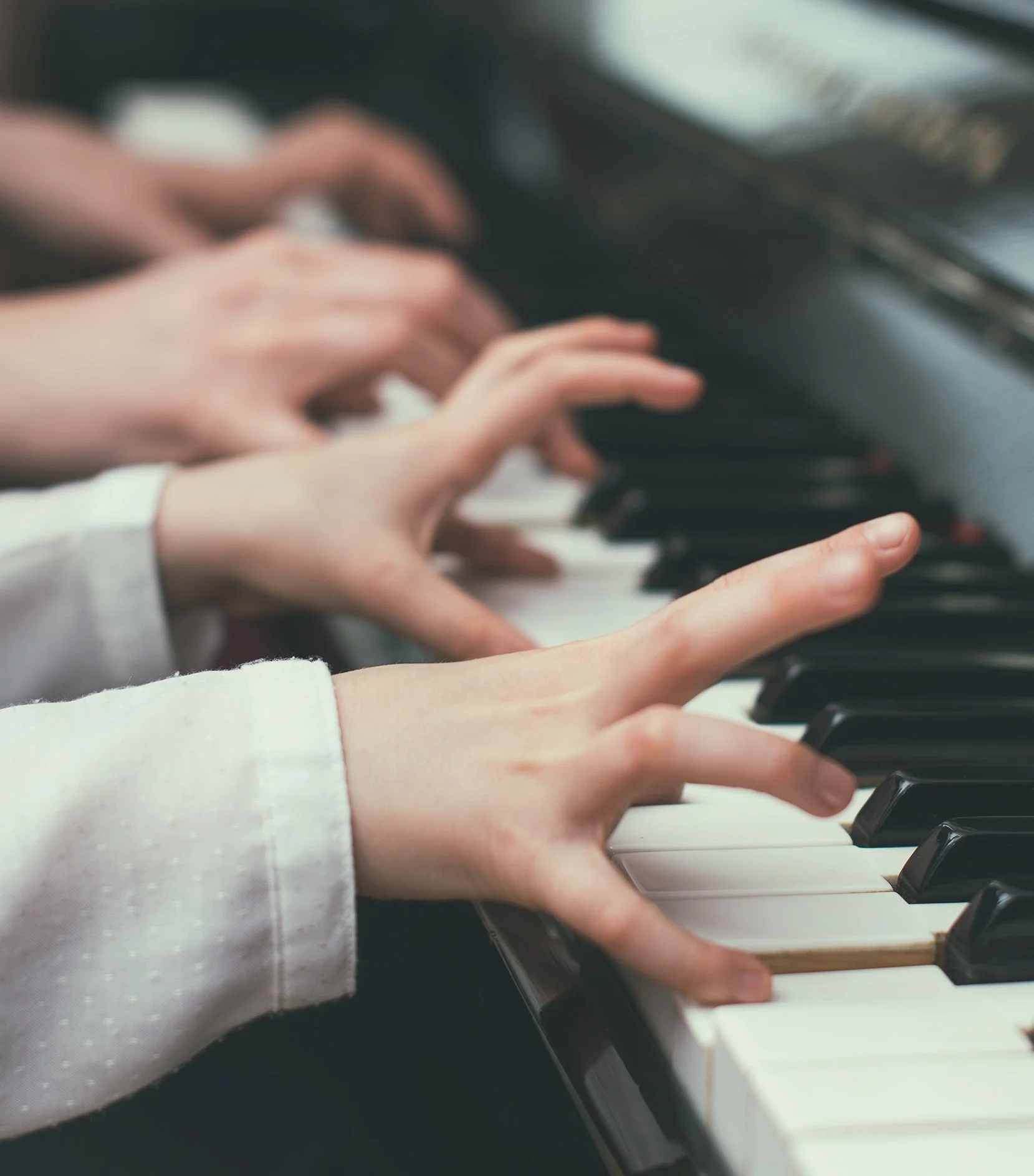 Two pairs of hands playing a piano, with focus on the hands and the keys.