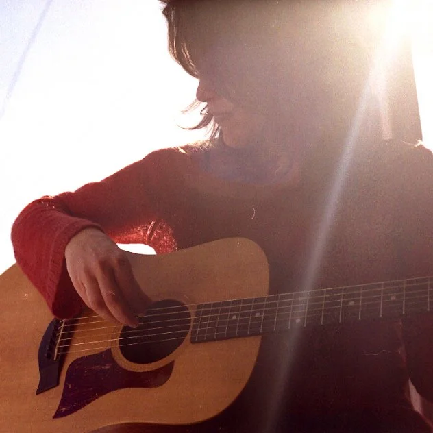 A woman sitting and playing an acoustic guitar, with sunlight shining in the background.