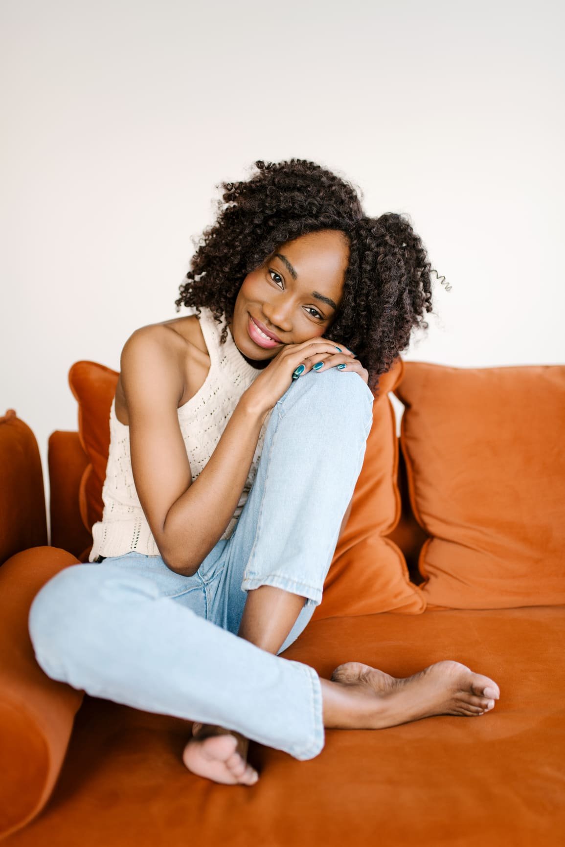 A black woman sitting on a sofa, her knee pulled up to her chest, she leans her head on her hand, on top of her knee