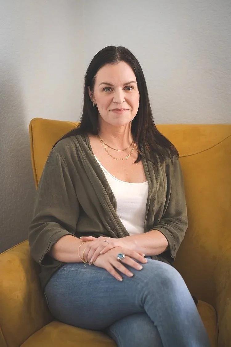 Carrie Hedinger sits in a yellow chair with her legs crossed, her hands stacked on her lap, and she smiles at the camera