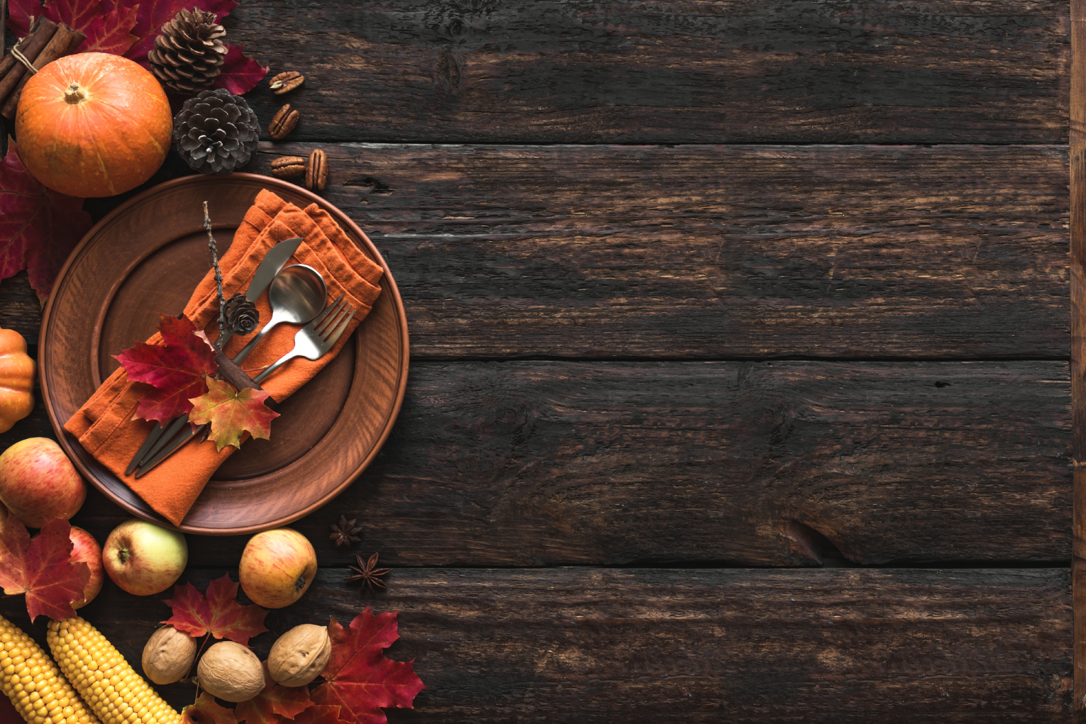 Thanksgiving table setting with autumn leaves, mini pumpkins, apples, corn, pine cones, walnuts, and silverware on an orange napkin on a wooden background.