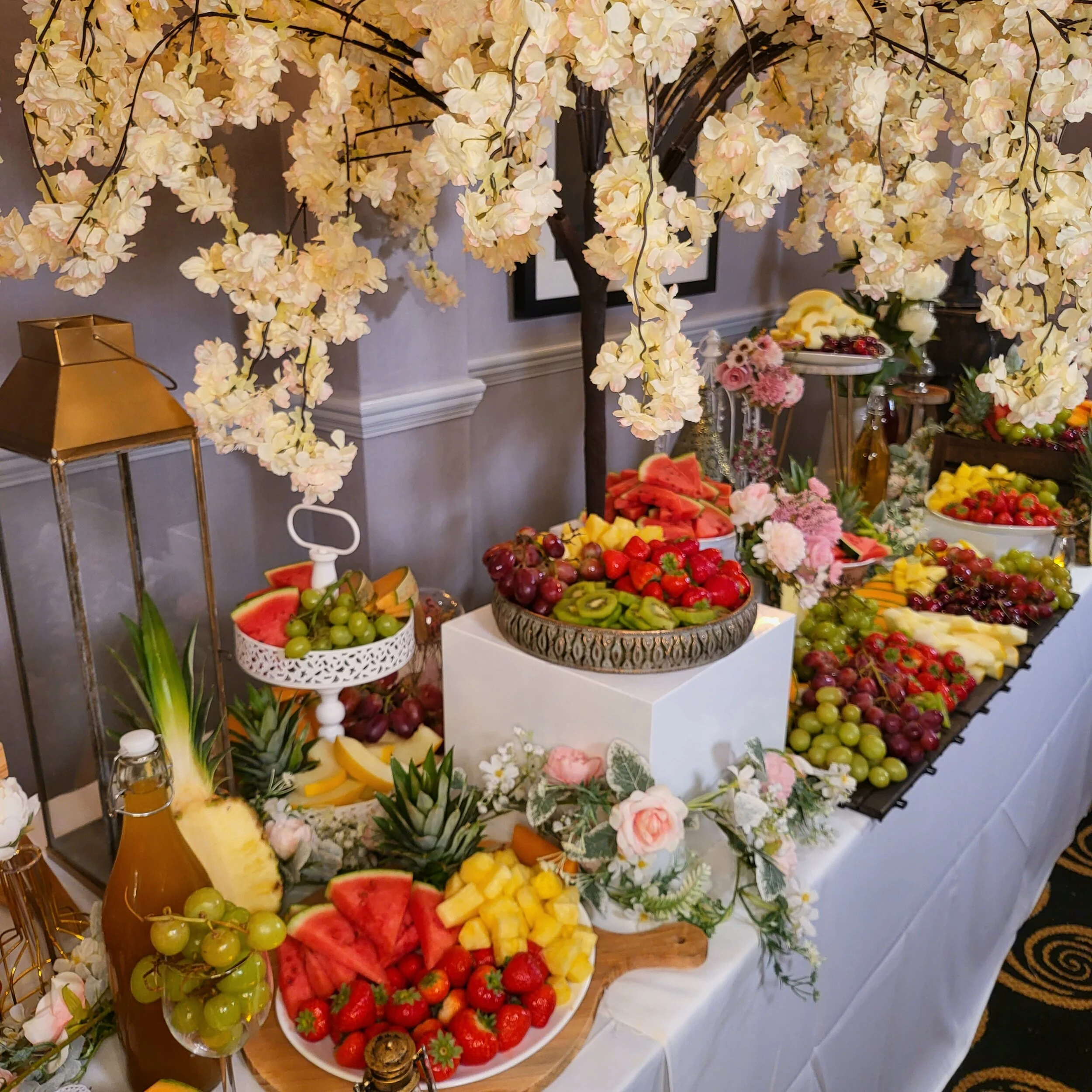 A dessert table with an abundance of fresh fruit, decorated with pink and white flowers and a flowering tree overhead.