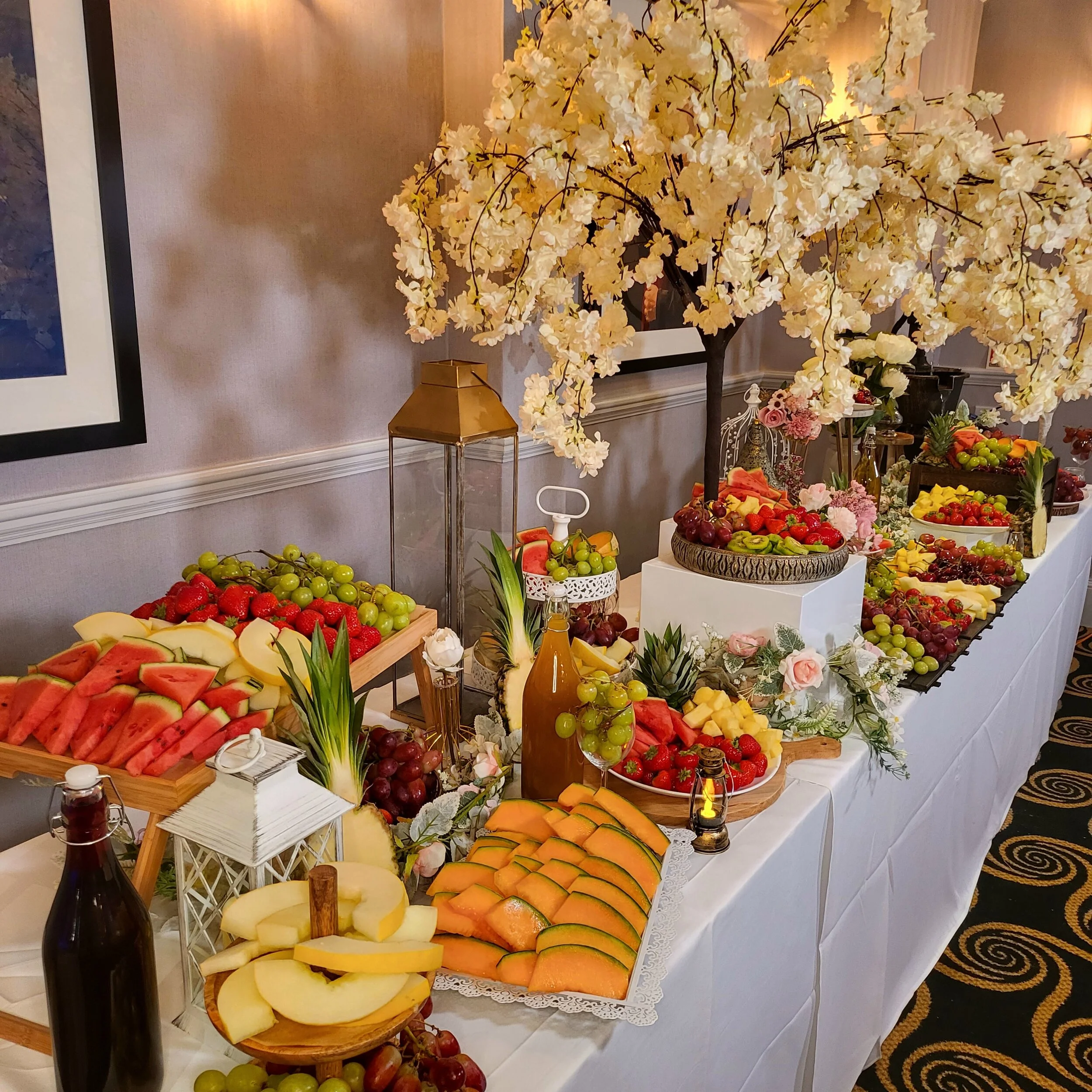 A fruit display table with various sliced and whole fruits, including watermelon, cantaloupe, honeydew, grapes, strawberries, pineapple, and cherries, decorated with a white floral tree and some lanterns.