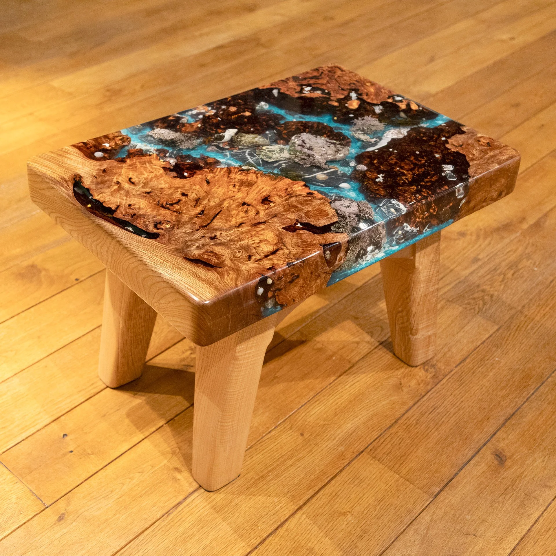 A small wooden table with a resin-embedded wood top, featuring a mix of natural wood grain and blue, brown, and black resin patterns, placed on a hardwood floor.