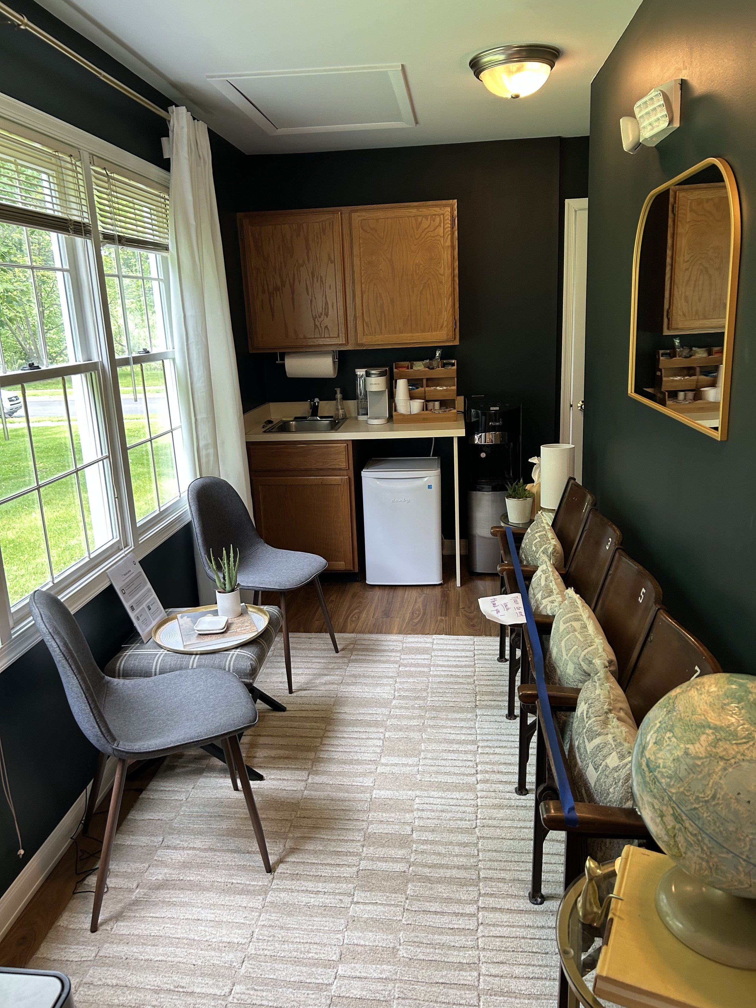 A small kitchenette and seating area with two chairs and a bench. There are large windows with white curtains on the left, a small counter with a sink, mini fridge, coffee machine, paper towel holder, and some shelves in the back. The walls are dark