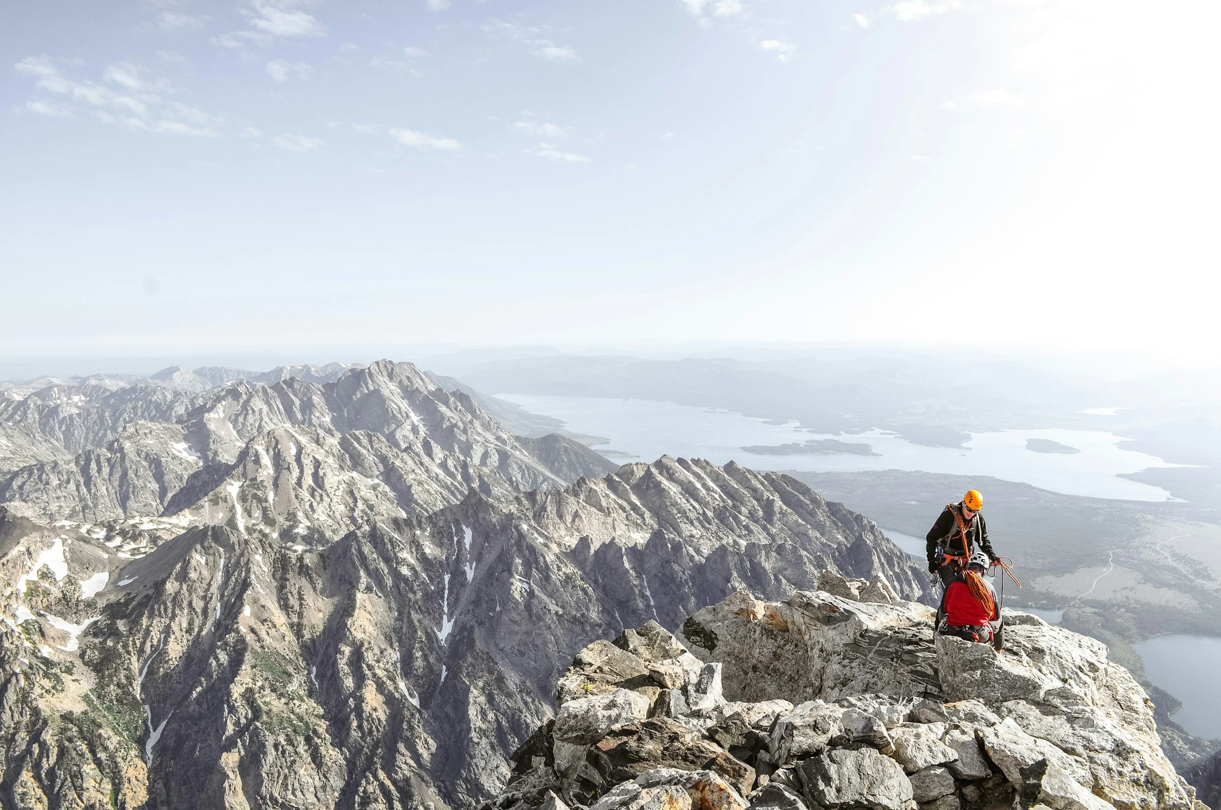 Two climbers at the summit of a mountain, with a vast mountain range and lake in the background.