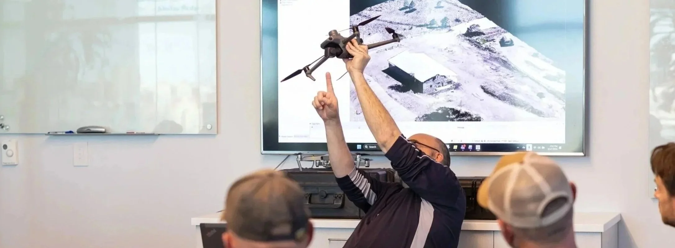 Instructor demonstrates a drone to students in a classroom with a large display screen showing a snowy landscape.