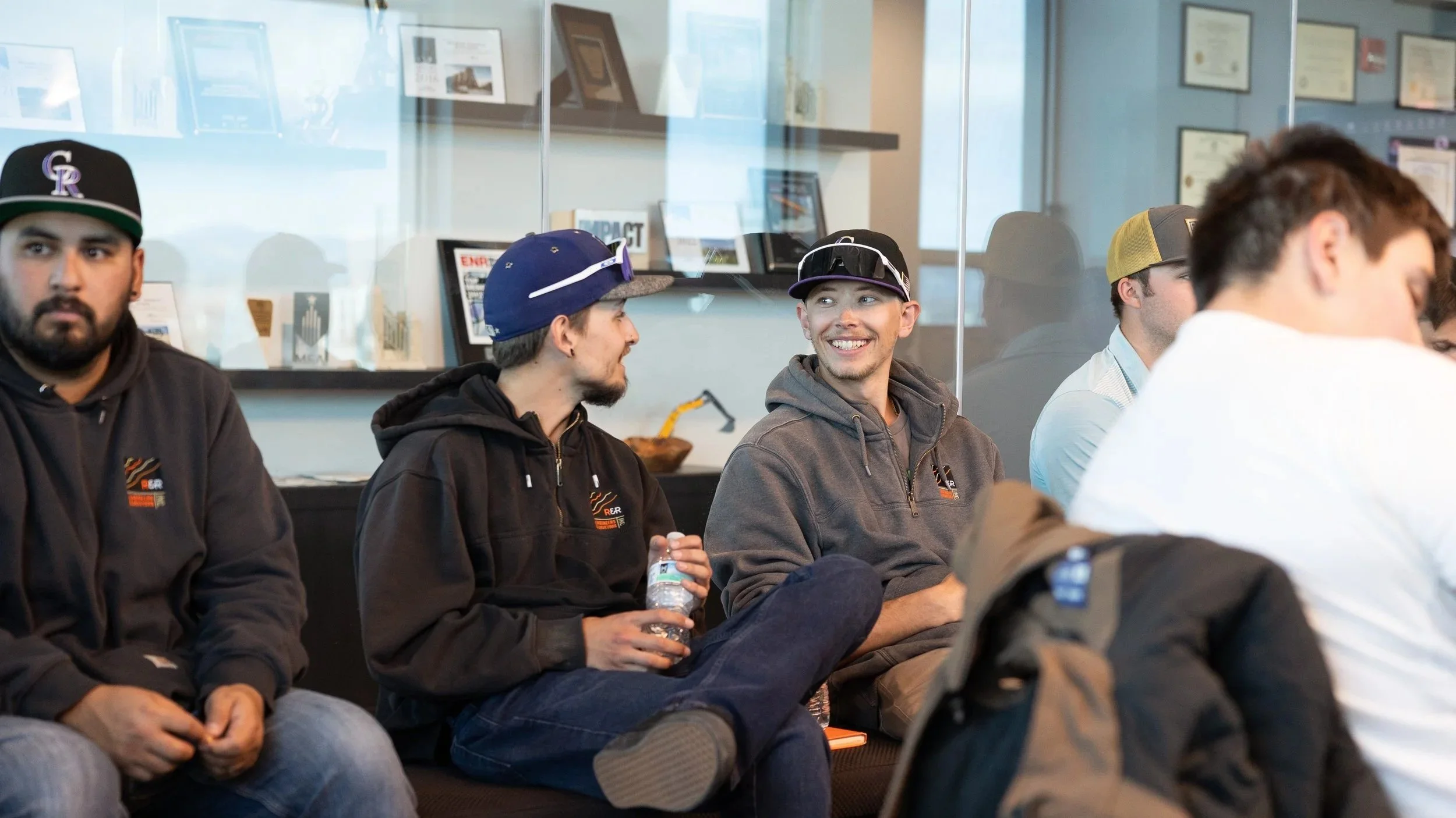 Group of young men sitting and talking in a waiting room, one holding a water bottle, some wearing baseball caps and hoodies.