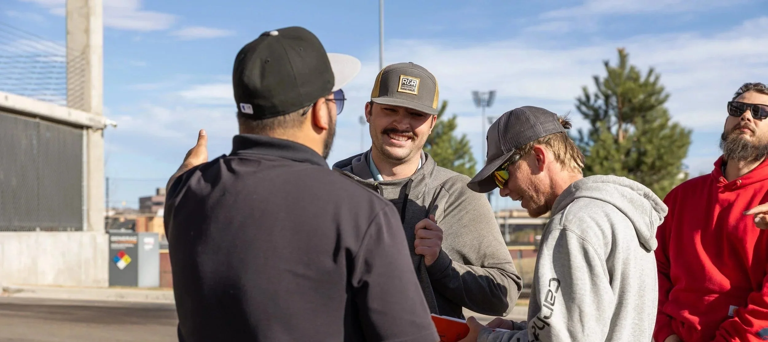 Four men talking outdoors on a sunny day, one wearing a black jacket and cap, another with a gray hoodie and a gray cap, a third with sunglasses and a red hoodie, and the fourth with a baseball cap, smiling and engaged in conversation.