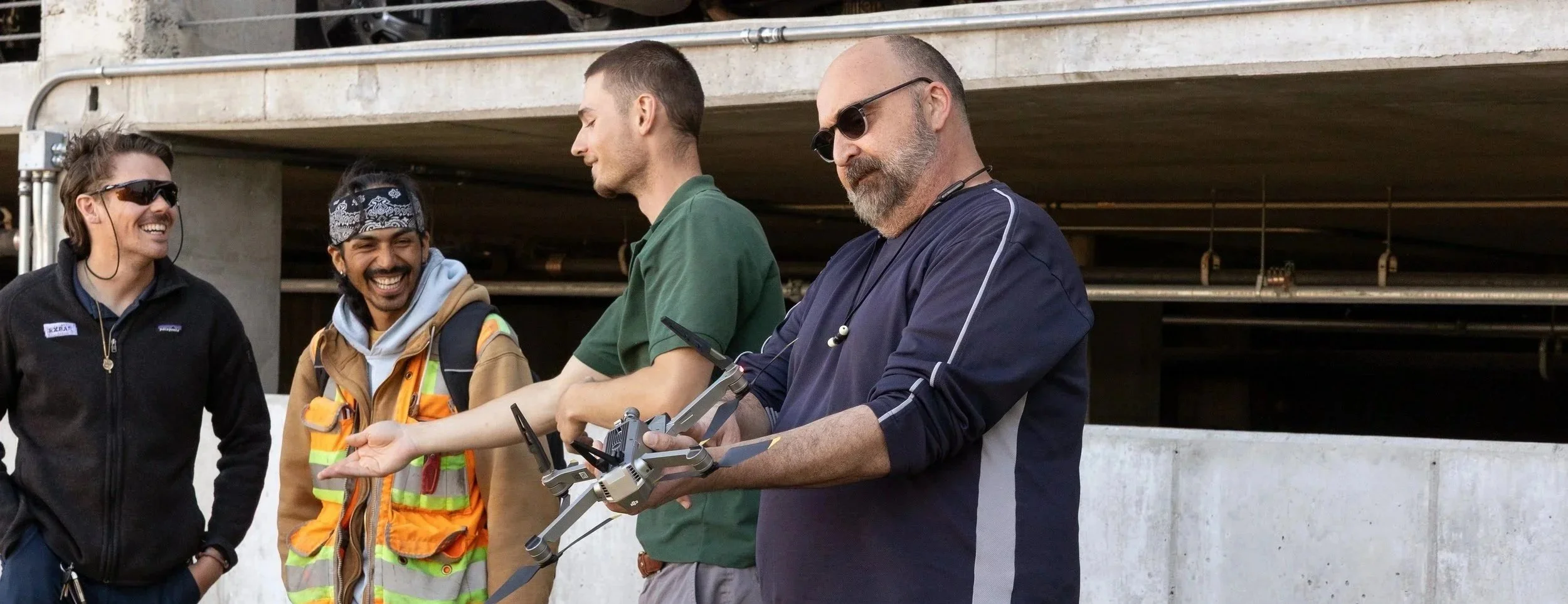 Four men standing outdoors near a parking garage, one man holding a drone, all smiling and engaging in conversation.