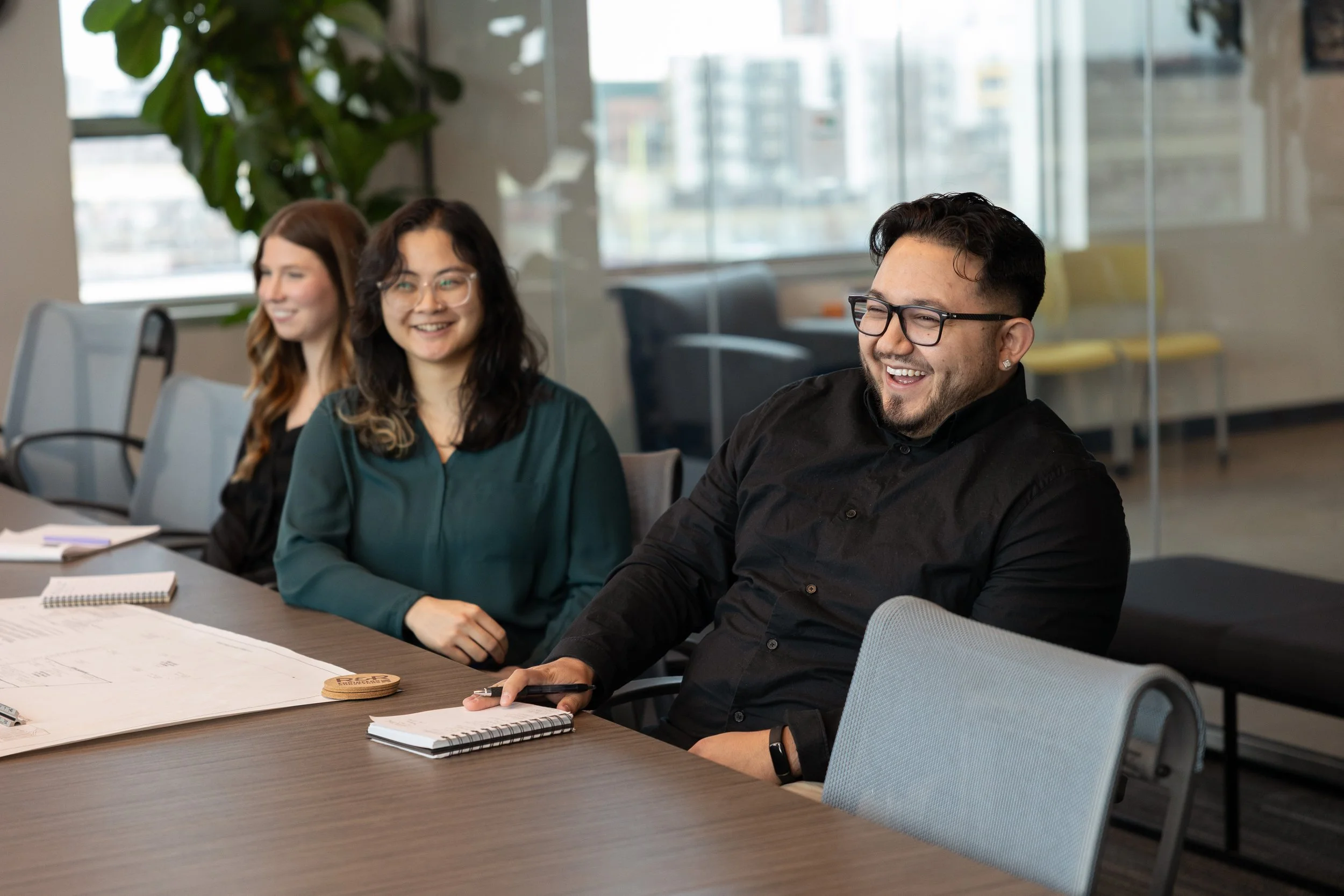 Four people sitting at a table in a modern office, talking and smiling. There are notebooks, papers, and a phone on the table.