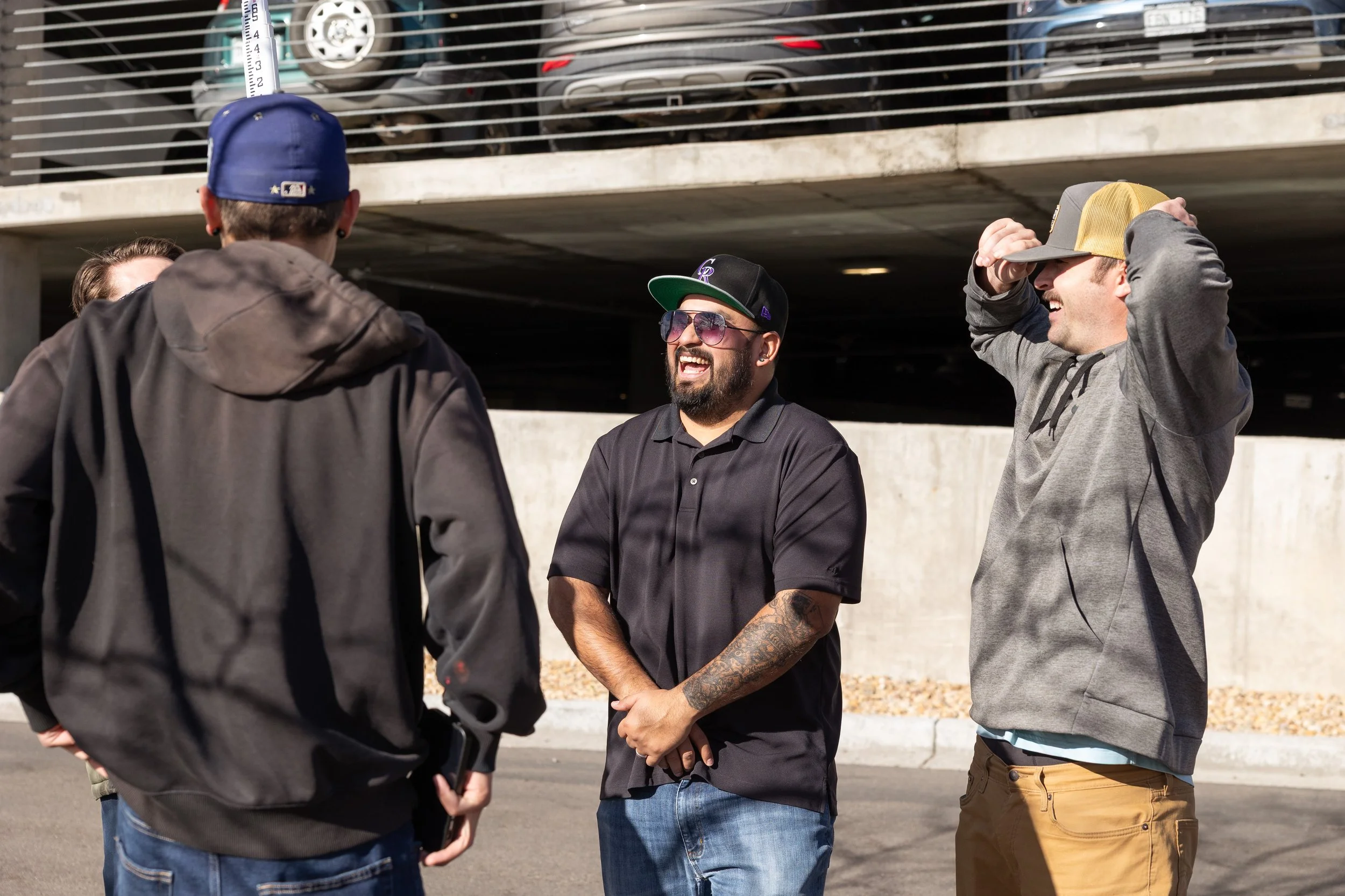 Four men standing in a parking lot having a conversation. One man in a black shirt, sunglasses, and a cap is smiling and clasping his hands in front of him. Another man in a gray hoodie and baseball cap is laughing, with his hand on his head. Two other men are partially visible from behind, facing the group.