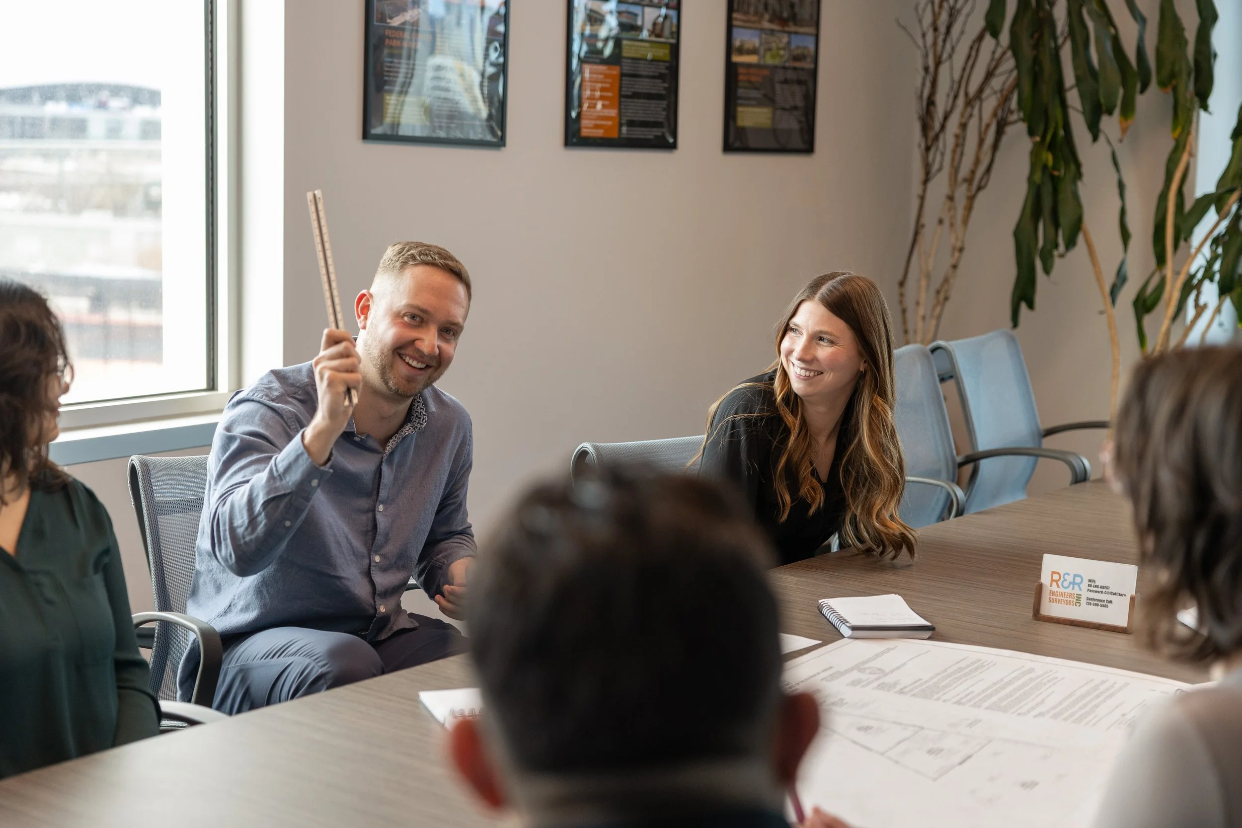 People in a business meeting in a conference room, with one man smiling and holding a pen, and women sitting around the table, some with papers and notebooks, with posters on the wall and a window showing an urban view.