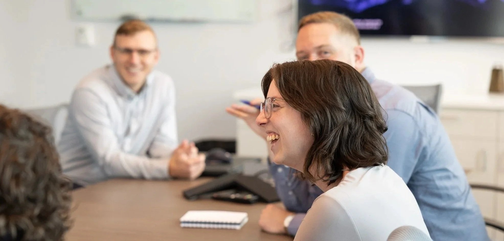 A group of four people in a meeting room, smiling and engaging in discussion, with notebooks and electronic devices on the table.
