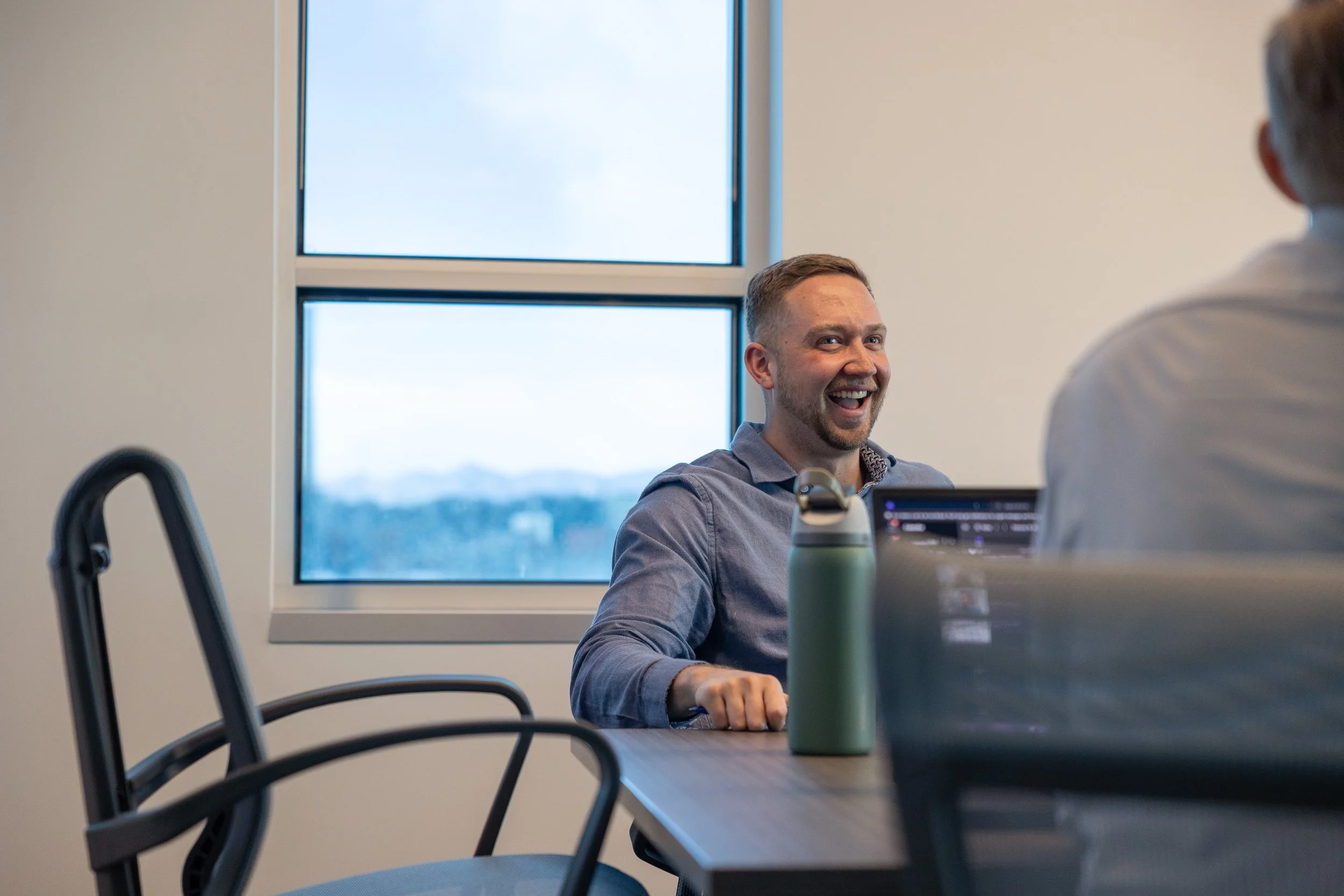 A man with short hair and a beard smiling and talking to someone else in an office setting, with a large window showing a cloudy sky in the background.