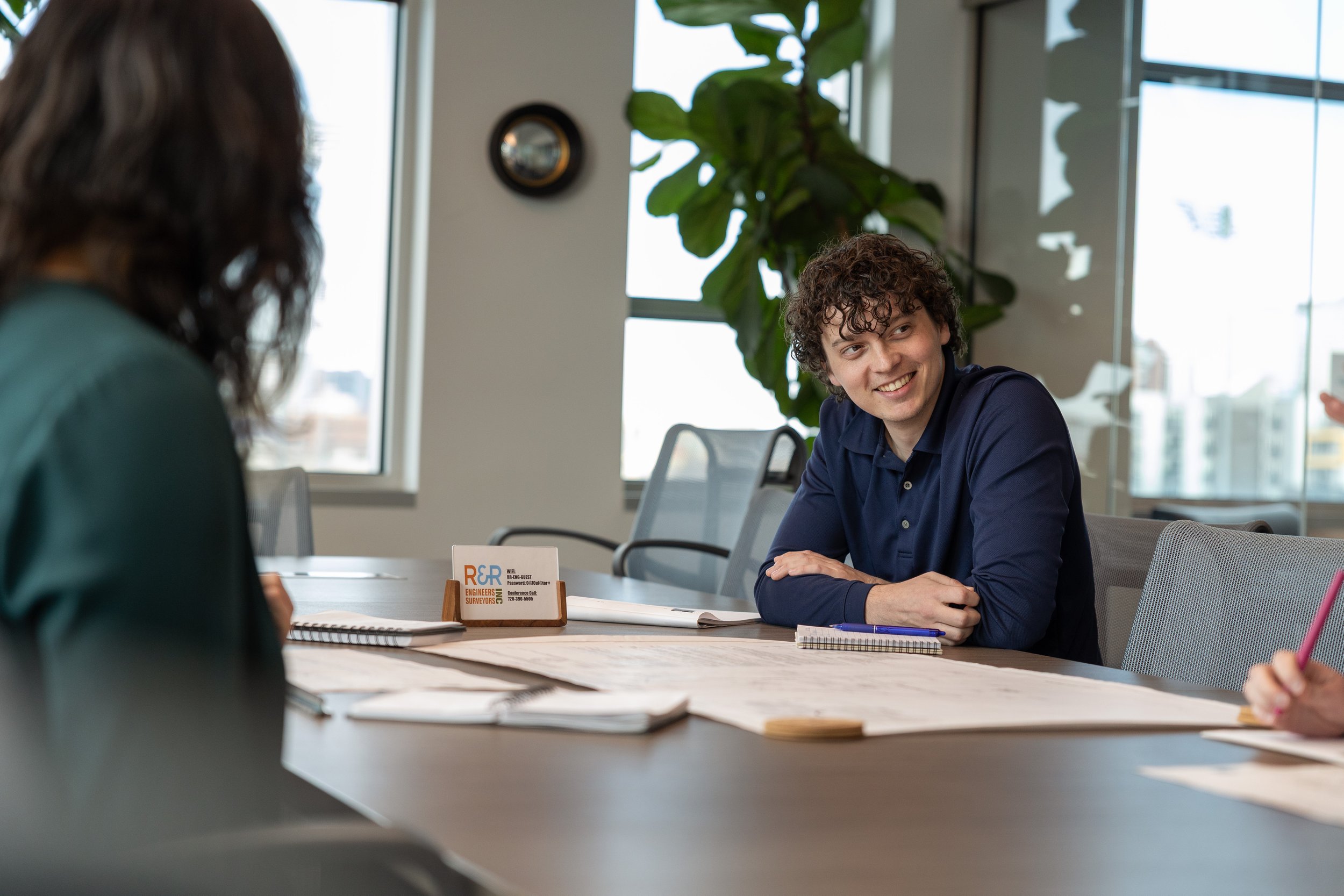 A young man with curly hair smiling while speaking with a woman who is partially visible, in a modern office during a meeting.