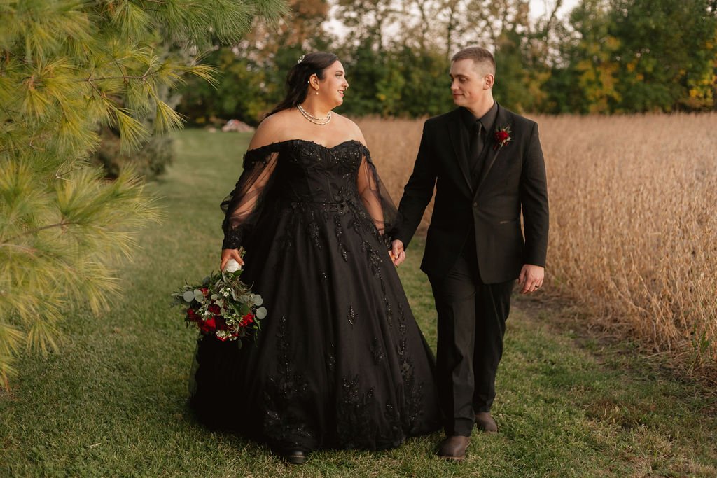 Bride and groom, both dressed in black, walking together along the rustic farm field at Country Coach House wedding venue in Burlington, IL.