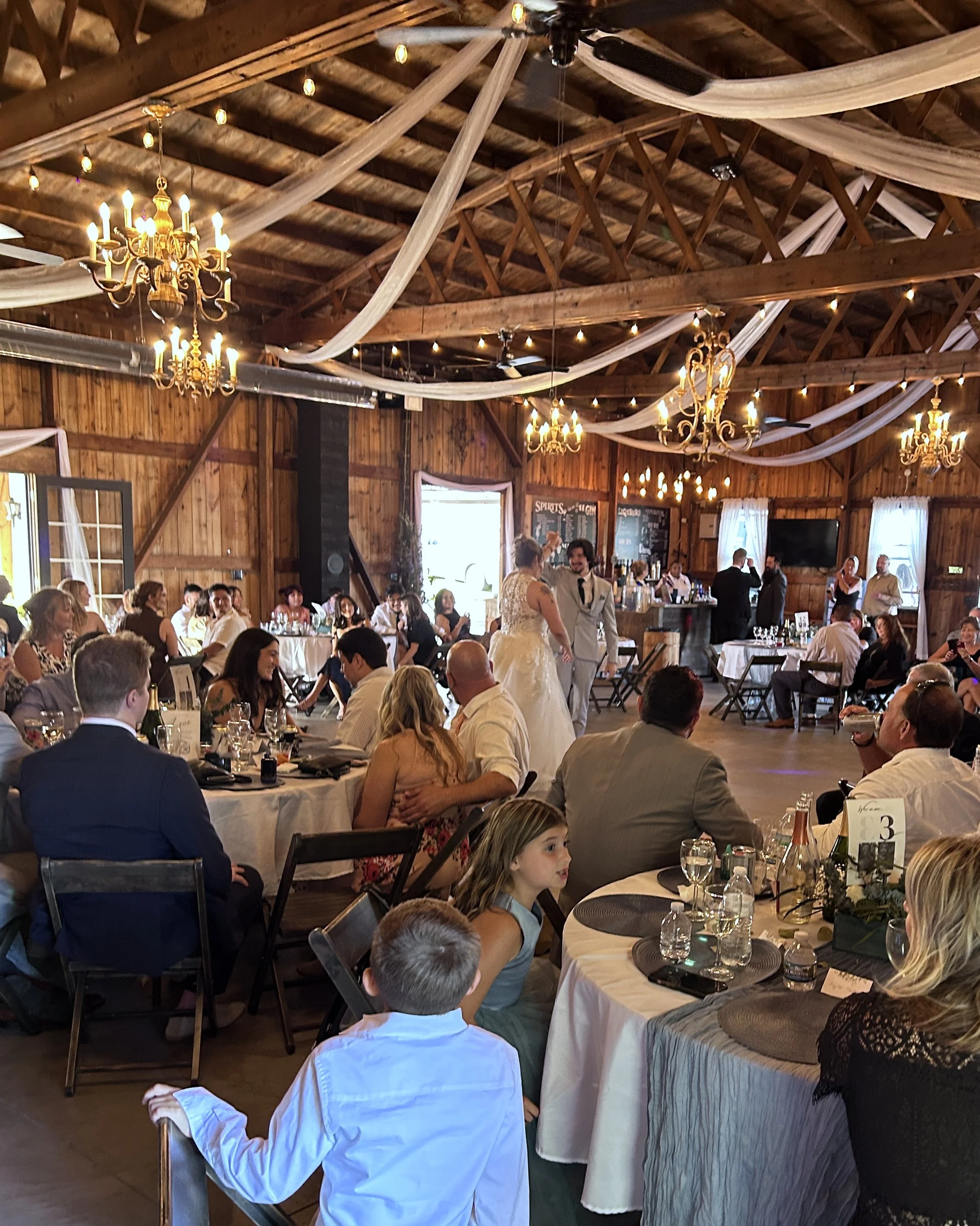 Guests seated inside the reception space at Country Coach House as the bride and groom are introduced. the image shoes the arched beams and chandeliers that add to the elegance of a rustic venue.