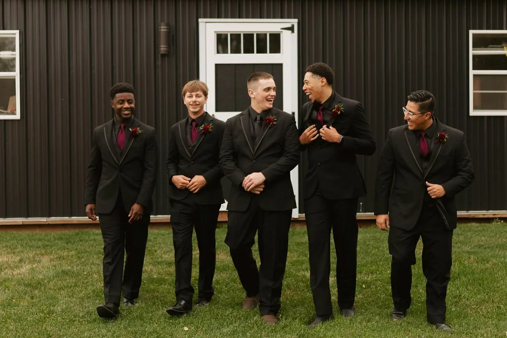 The groom and groomsmen, dressed in black with deep red ties and boutonnieres, share a relaxed moment with the black shed backdrop at Country Coach House in Burlington, Illinois.