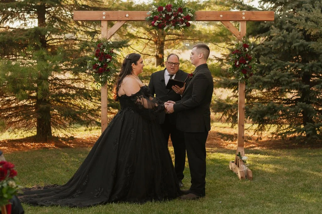 Bride and groom with backdrop of pine trees and red roses, exchanging vows during their outdoor wedding ceremoy at Country Coach House in Burlington, IL