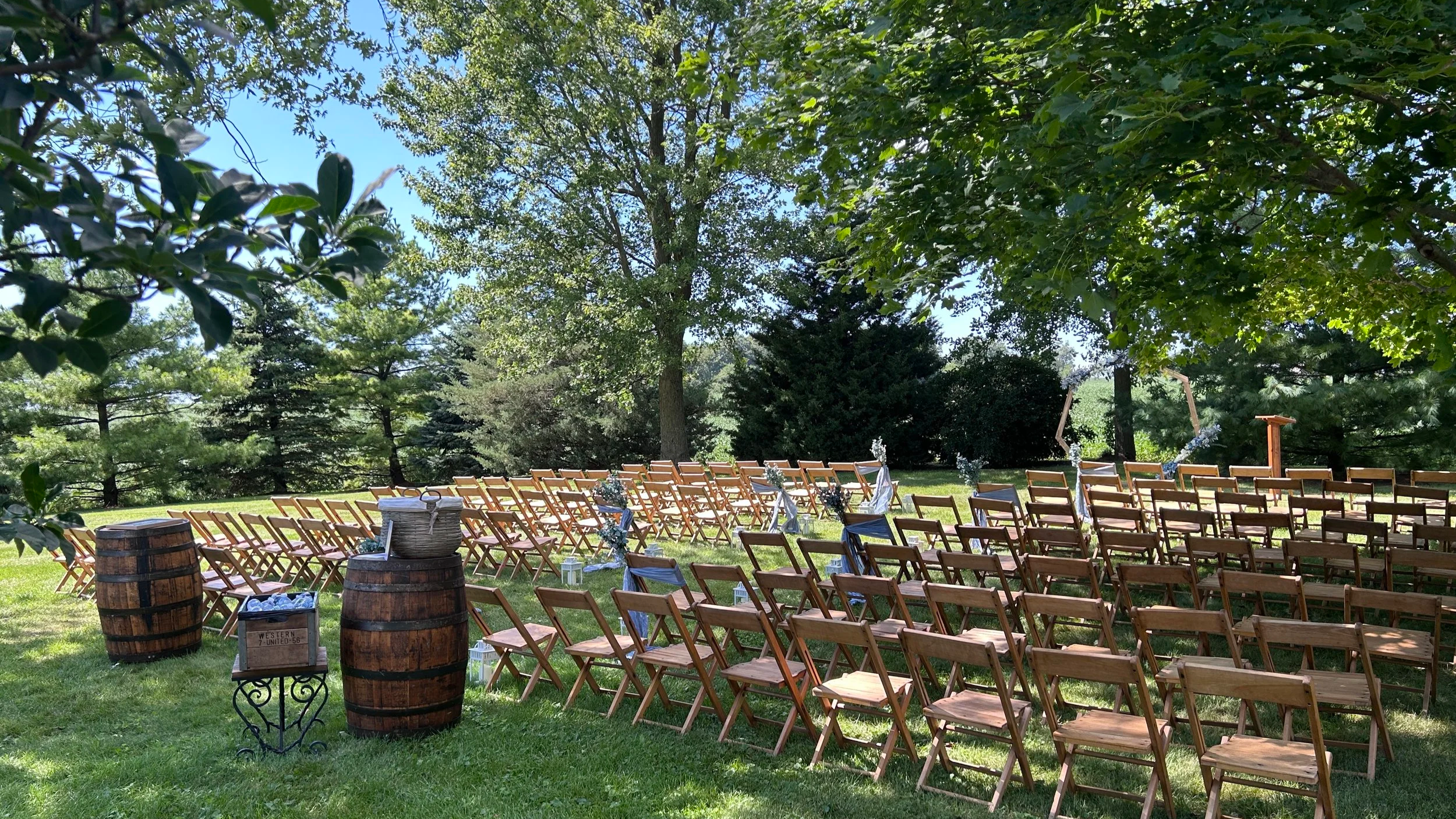 Ceremony chairs in neat rows with two barrels marking the aisle, water bottles in a rustic crate, and a blue floral heptagon arch at our Northern Illinois barn venue.