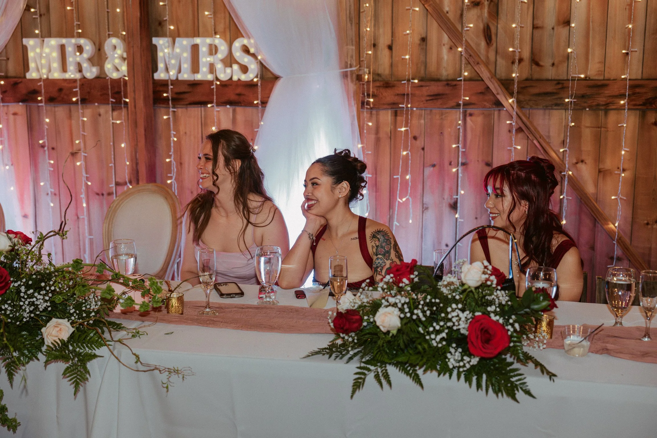 Rose colored uplights against the wood walls at Country Coach House, the head table with rose runners, red and white roses, on white linens with the warmth of gold votive  candles, and champaign filled glasses, and smiling bridesmaids.