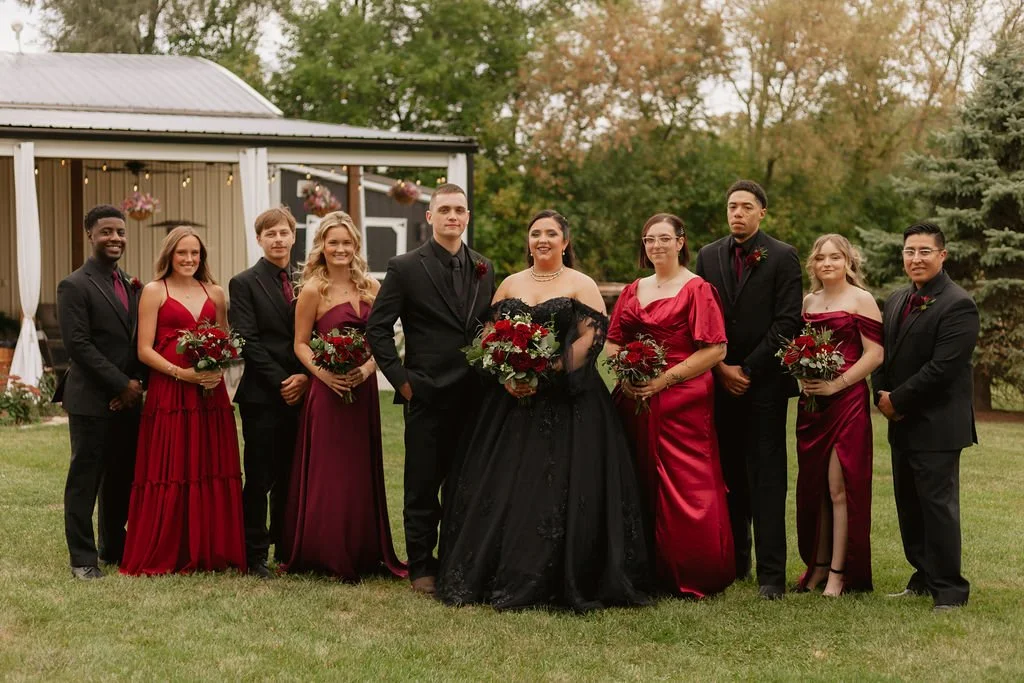 The wedding party dressed in black and red stands together with trees and the covered porch behind them at a Country Coach House wedding in Burlington, Illinois.