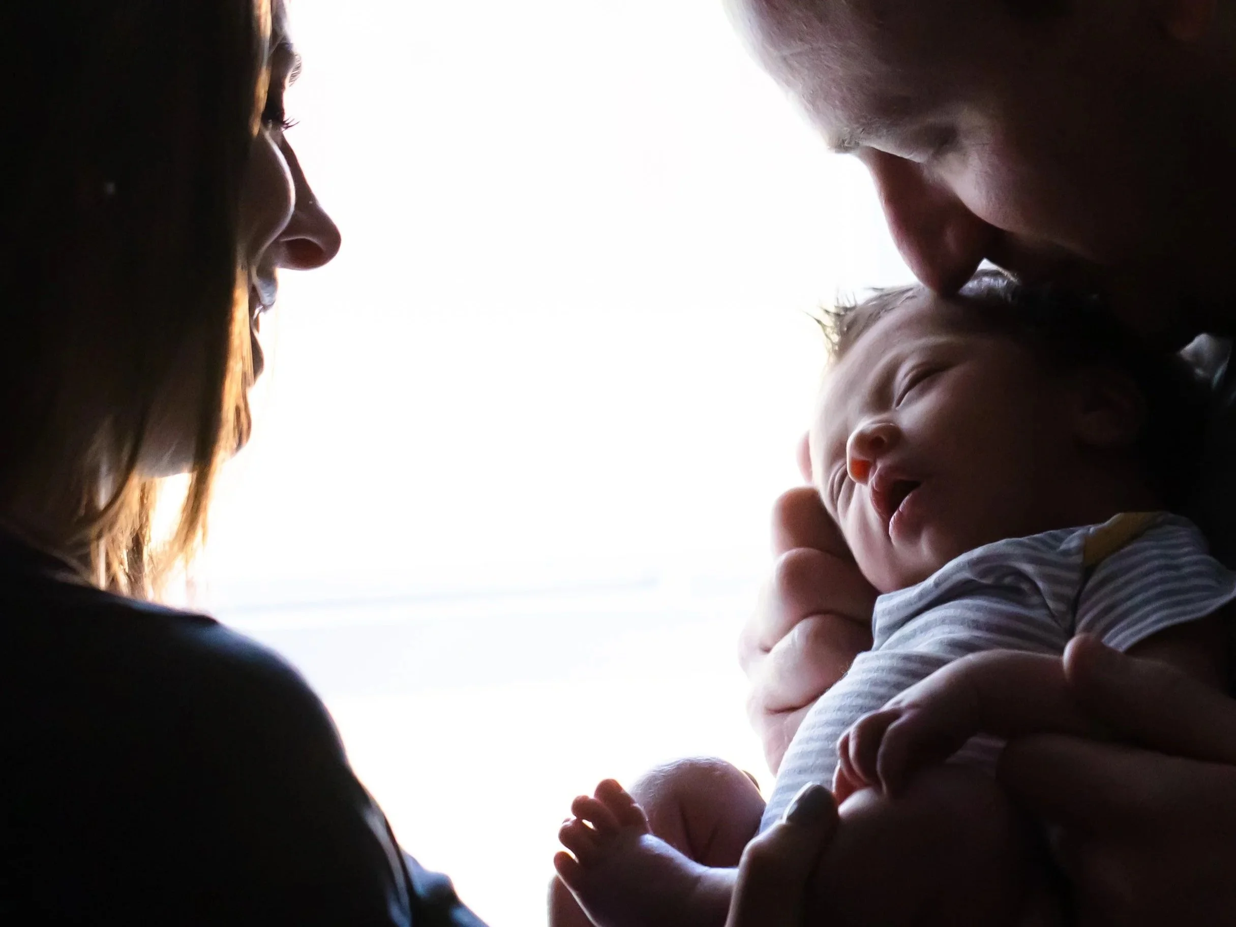 A woman, a man, and a baby are closely gathered, with the woman and man leaning in towards each other and the baby smiling and looking up. The photo is taken from below, with a bright light background.