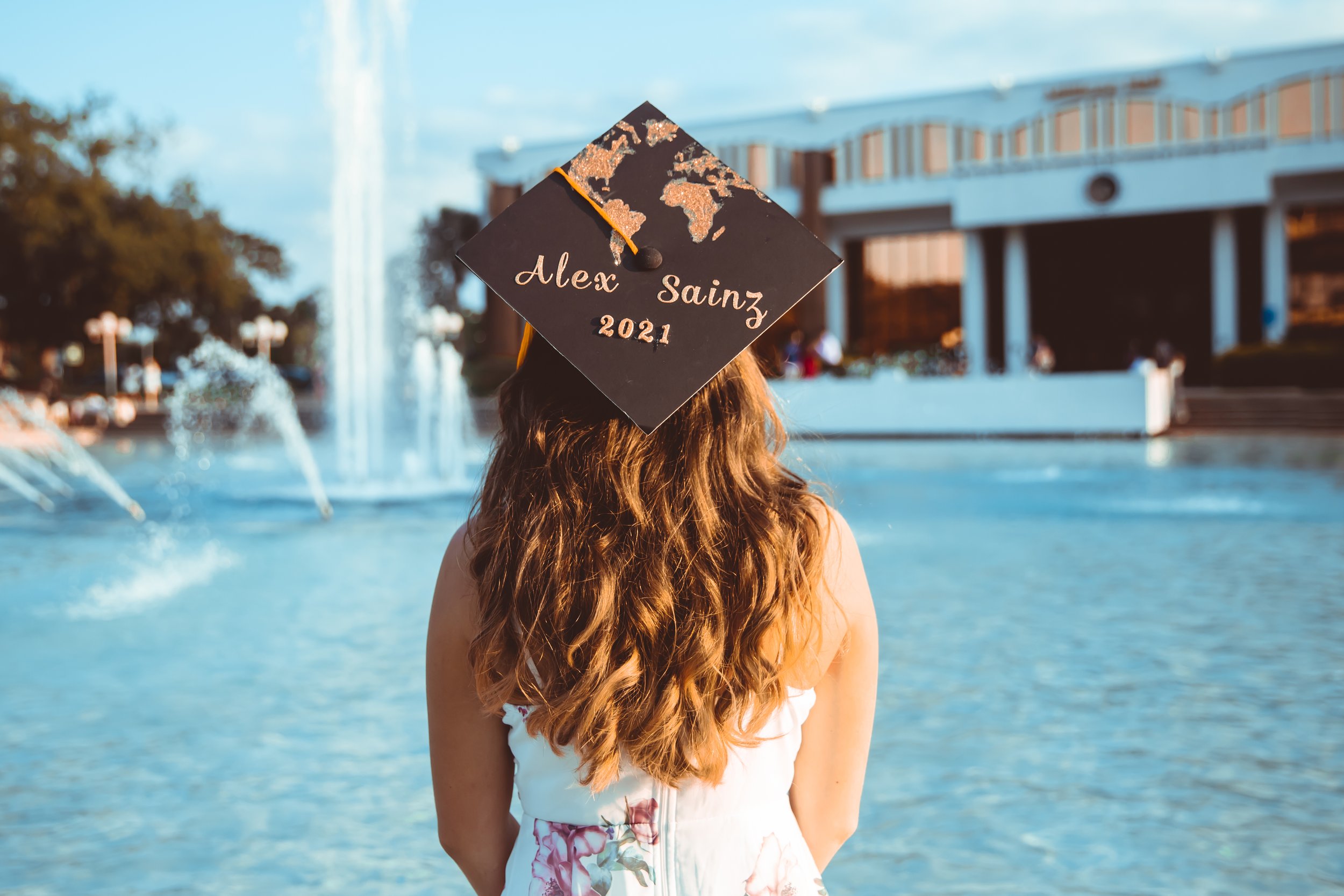 Person with long, wavy hair is standing near a fountain with jets of water, wearing a black graduation cap with a world map design and the name 'Alex Sainz 2021' written on it. The background features a large modern building with columns and glass, a
