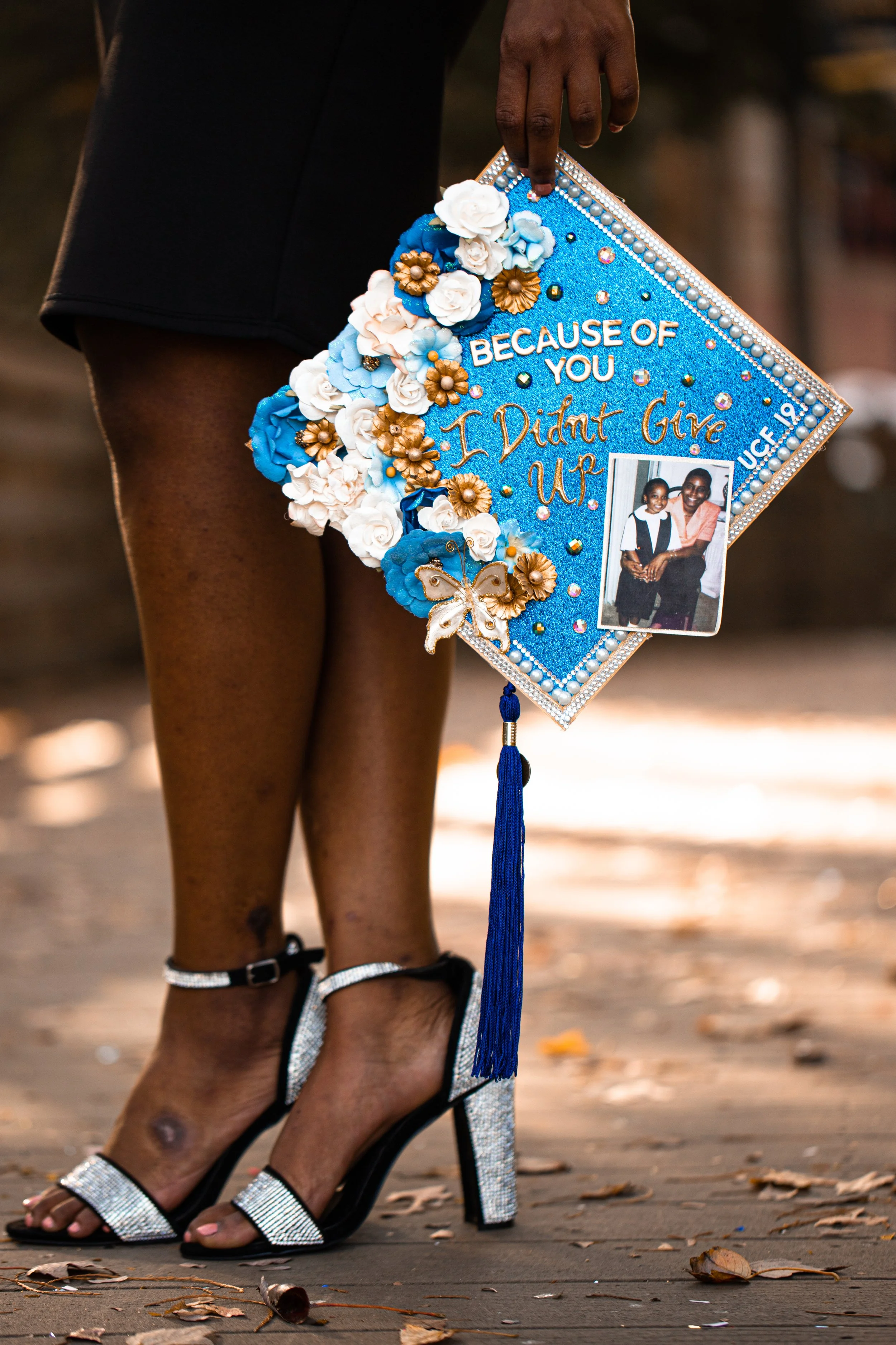 A decorated graduation cap with flowers, pearls, and rhinestones, held by a person standing outdoors on a wooden surface with fallen leaves, wearing black and silver high-heeled sandals. The cap has the message "Because of you I didn't give up" and a