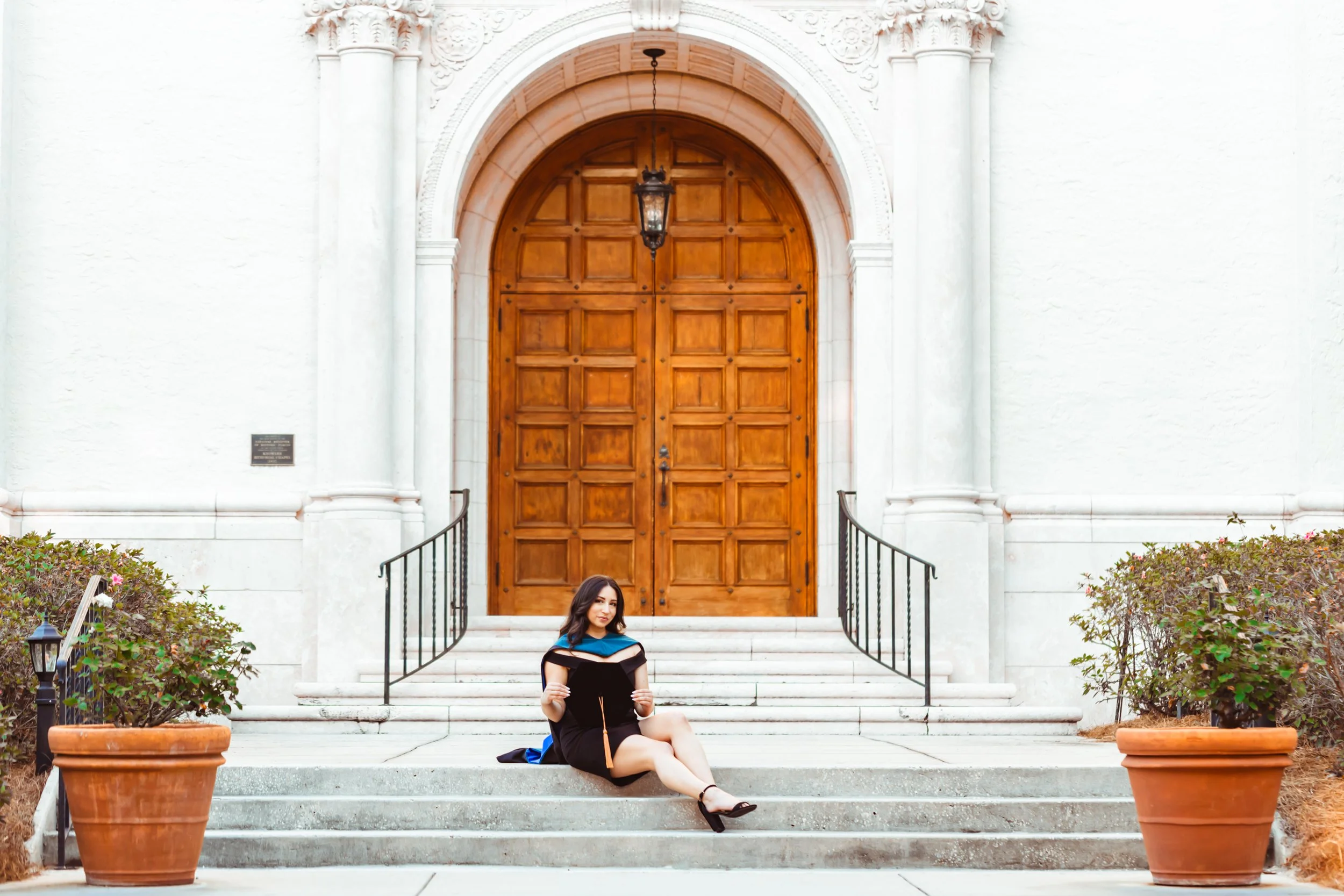 A woman sitting on steps in a graduation gown in front of a large wooden door with ornate white stone archway, flanked by potted plants. graduation photography session rollins college