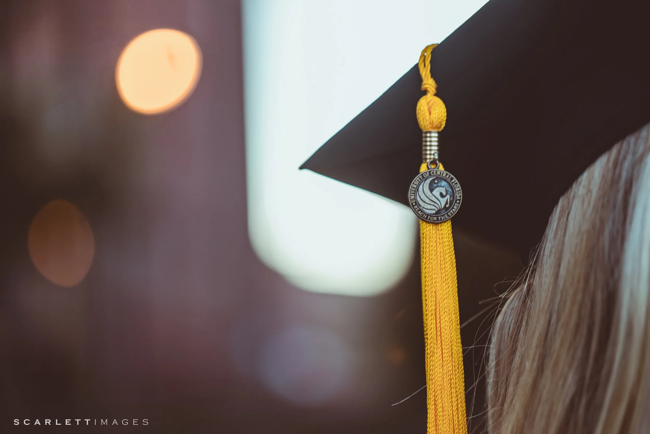 Close-up of a black graduation cap with a yellow tassel, showing a university badge. The background has blurred lights.