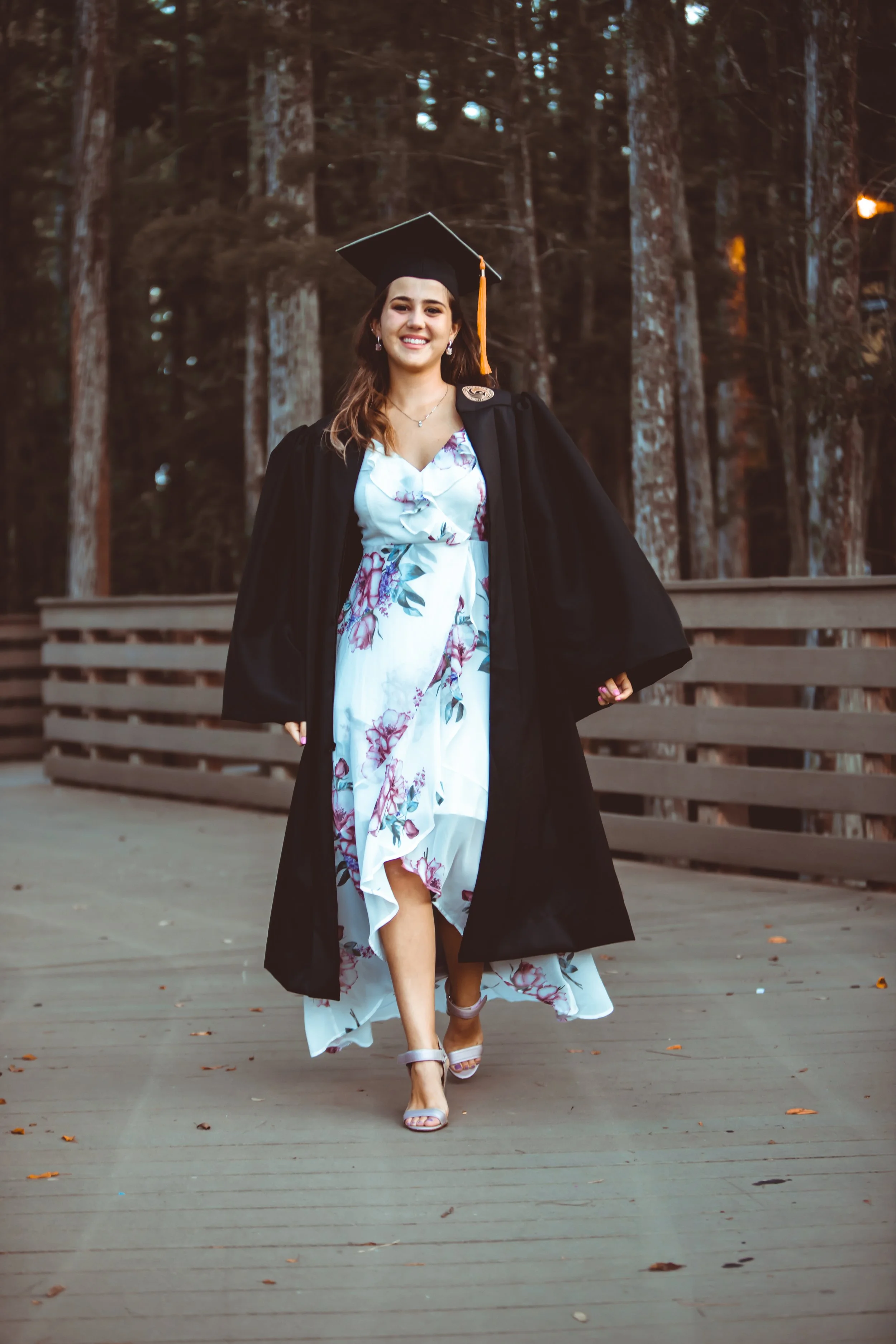 A young woman in a graduation gown and cap, smiling and walking on a wooden bridge in a forest setting, wearing a floral dress and high-heeled sandals.