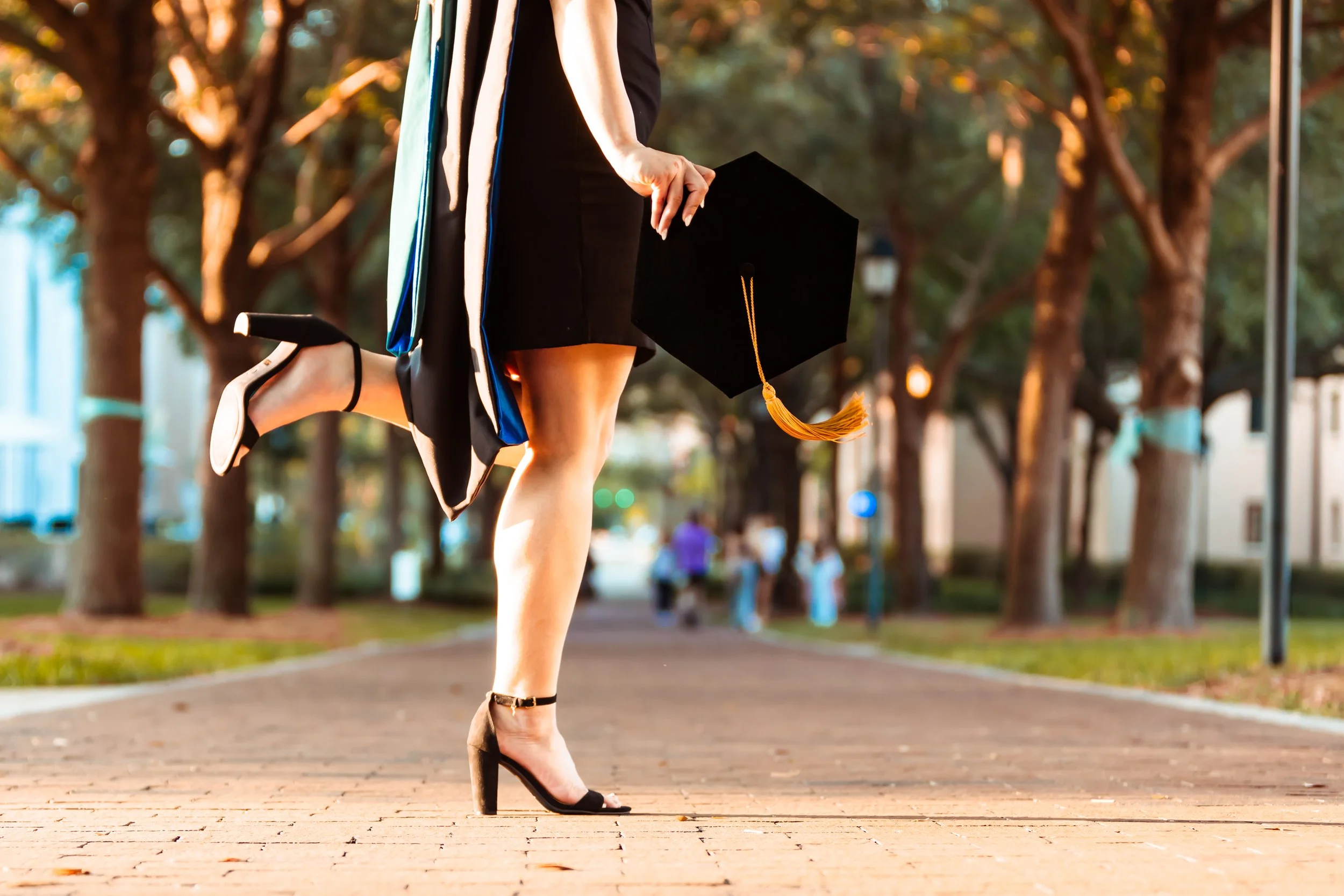 A woman in a black dress and high heels holding a graduation cap walking outdoors on a brick pathway with trees in the background.