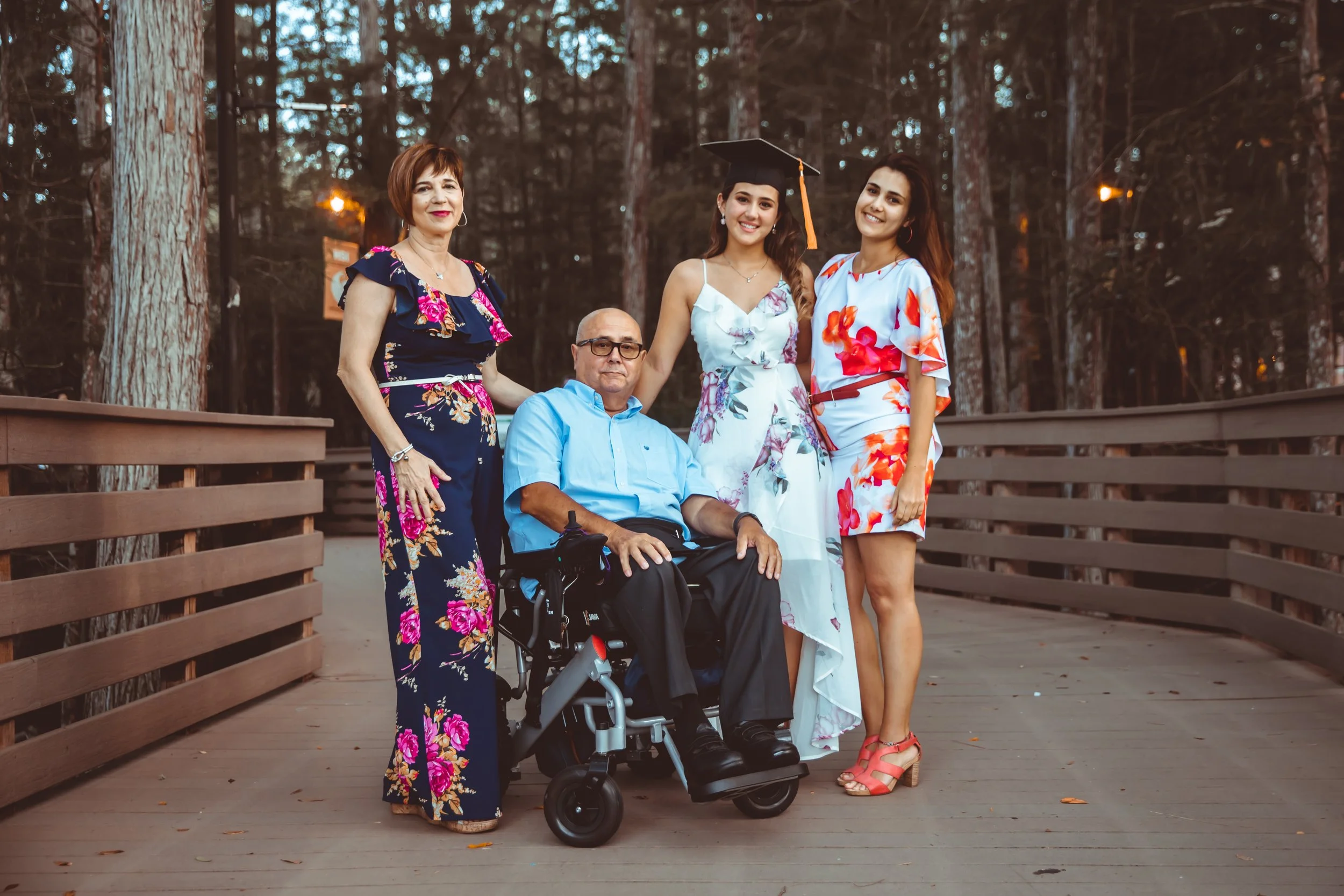 Group of five people outdoors on a wooden deck in a forest setting. Two women standing on either side of an elderly man in a wheelchair, with a young woman in a white dress and graduation cap in the middle. All are smiling.