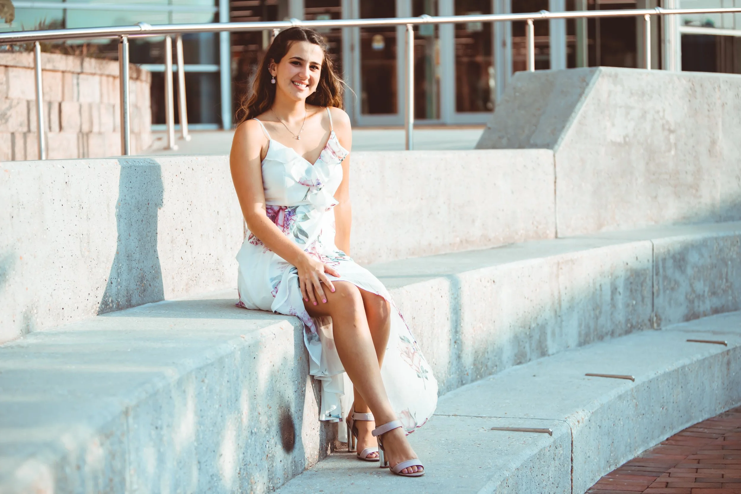 Smiling woman in a floral dress sitting on concrete steps outdoors.