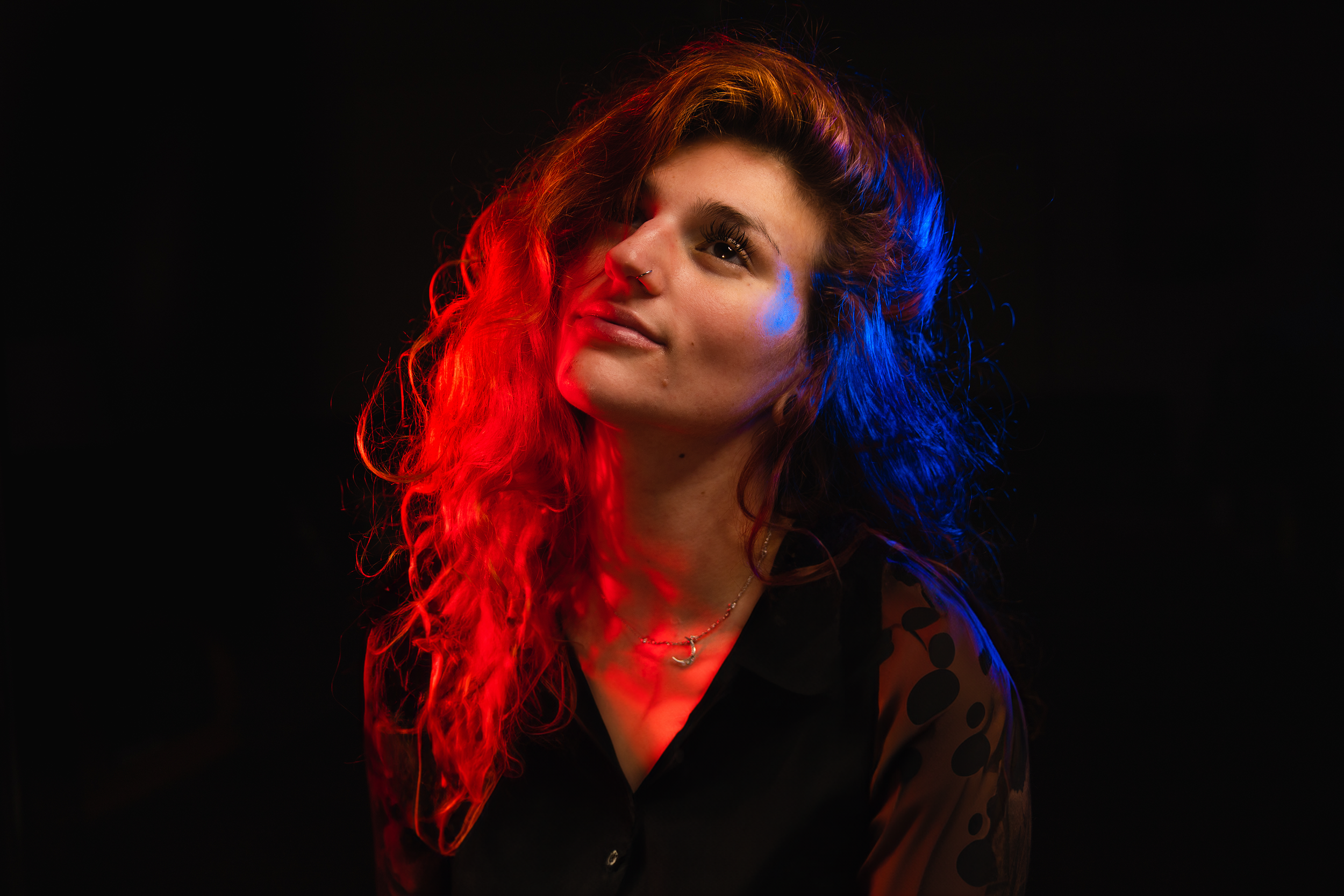 A woman with long curly hair illuminated with red and blue lighting, wearing a black top with Polka dots and a necklace, standing against a dark background.