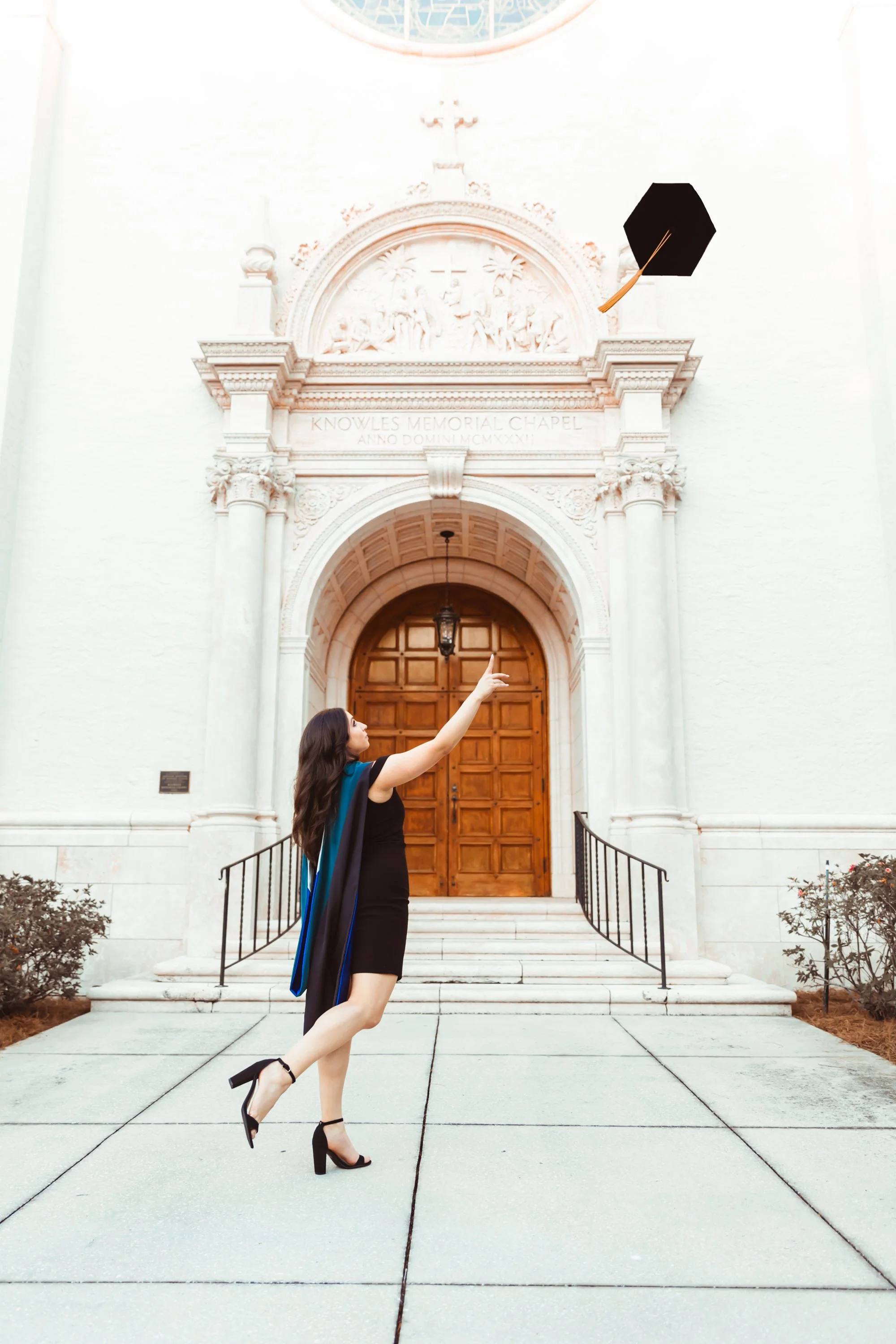 A woman in a black dress and high heels is standing on one leg in front of a church, pointing upward at a flying graduation cap with an tassel. The church has white walls, columns, a wooden door, and an arch with intricate carvings.