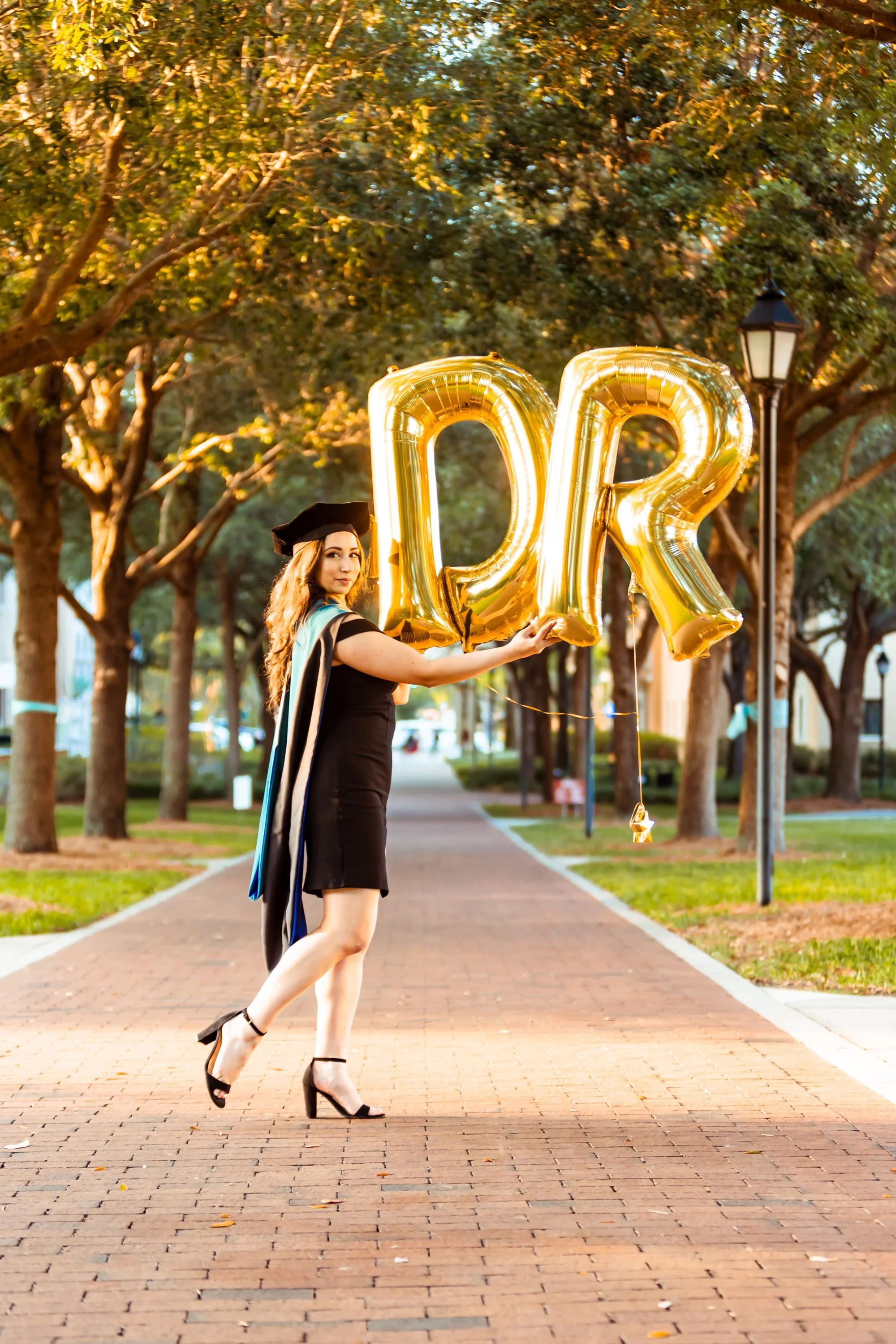 A woman in a graduation gown and cap holding gold foil balloons that spell 'GR'. She is wearing high heels and is standing on a brick pathway in a park-like area with trees and a streetlamp.