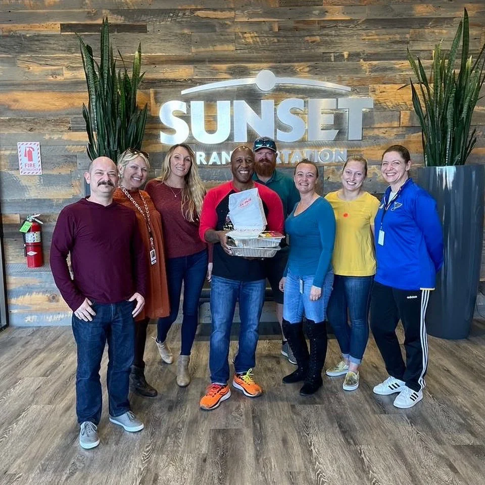 Steve of Steve's Hot Dogs stands with an employee group who has ordered catering for lunch with a bag of catering and two pans of warm food.
