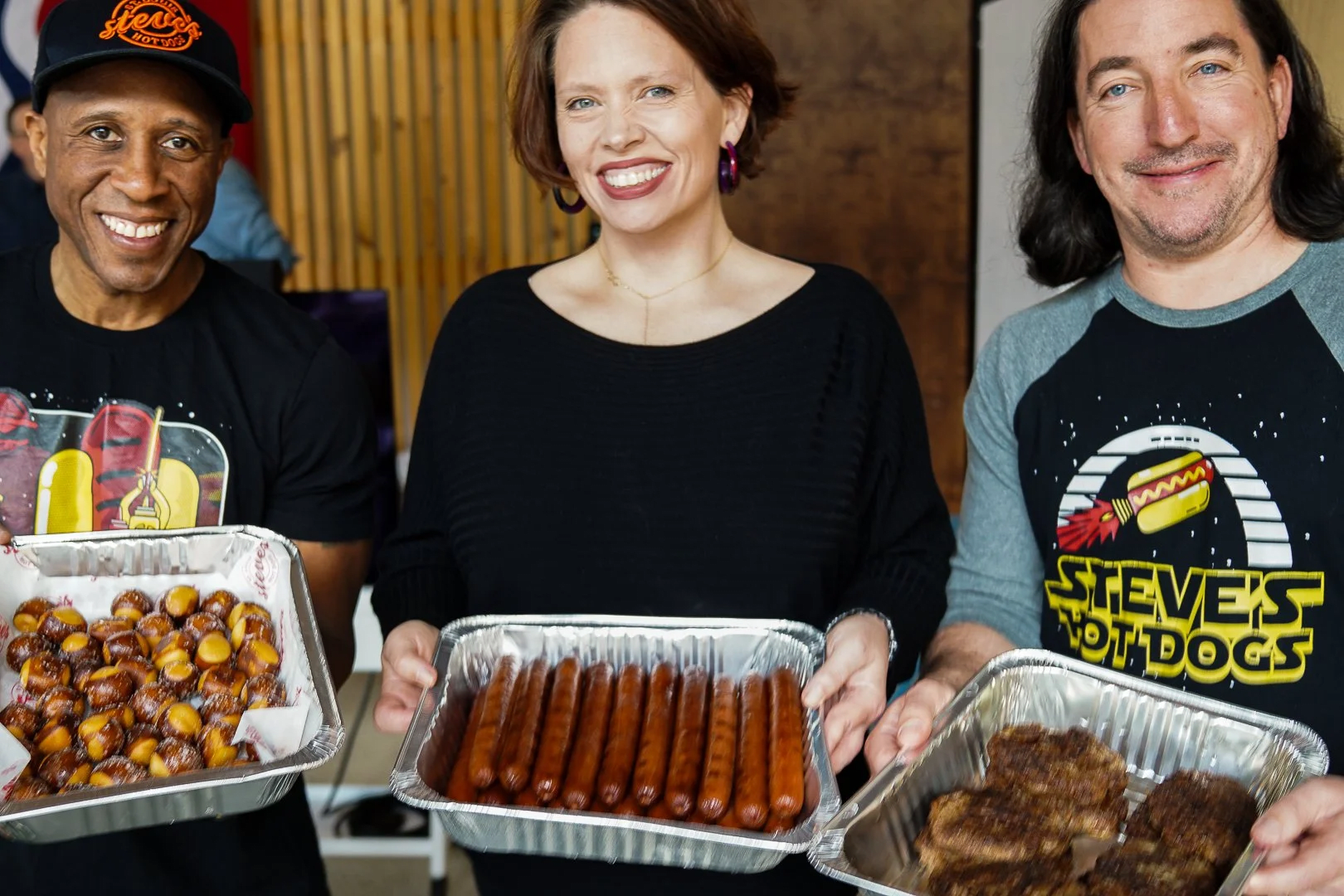 3 people holding 3 large pans of catering food - hot dogs, baked beans and pretzel bites