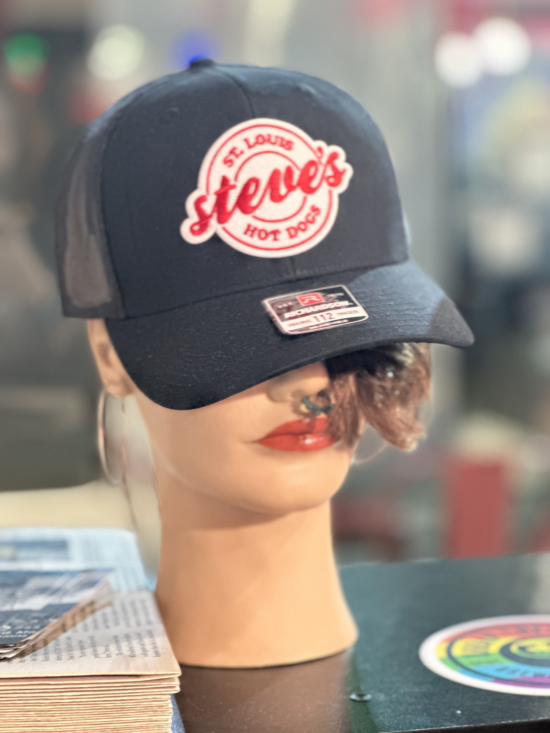 A black hat with a white and red patch with the Steve's Hot Dogs logo