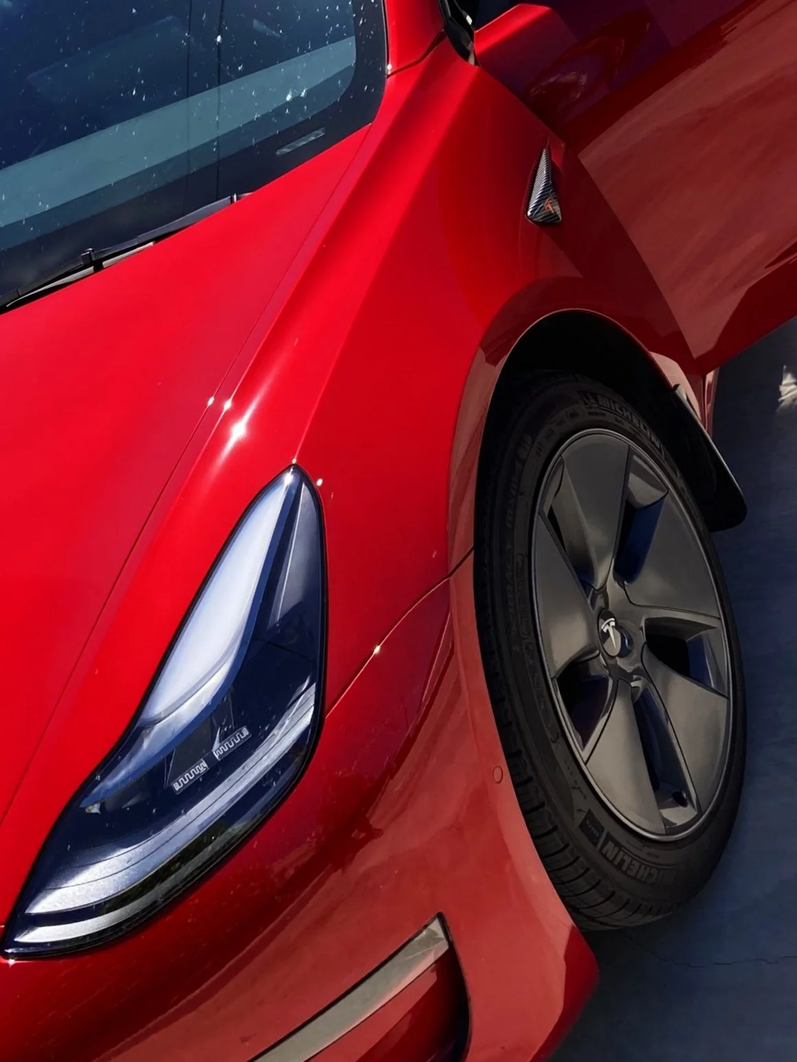 Close-up of a red Tesla Model 3, showing the front left headlight, wheel, and part of the fender.